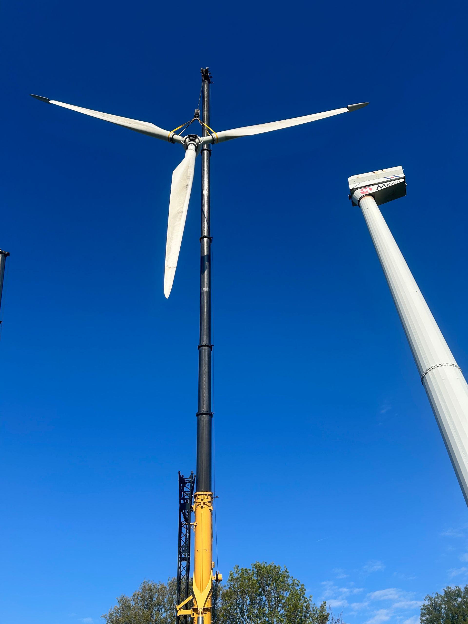 Two wind turbines against a clear blue sky, one partially assembled on a crane.