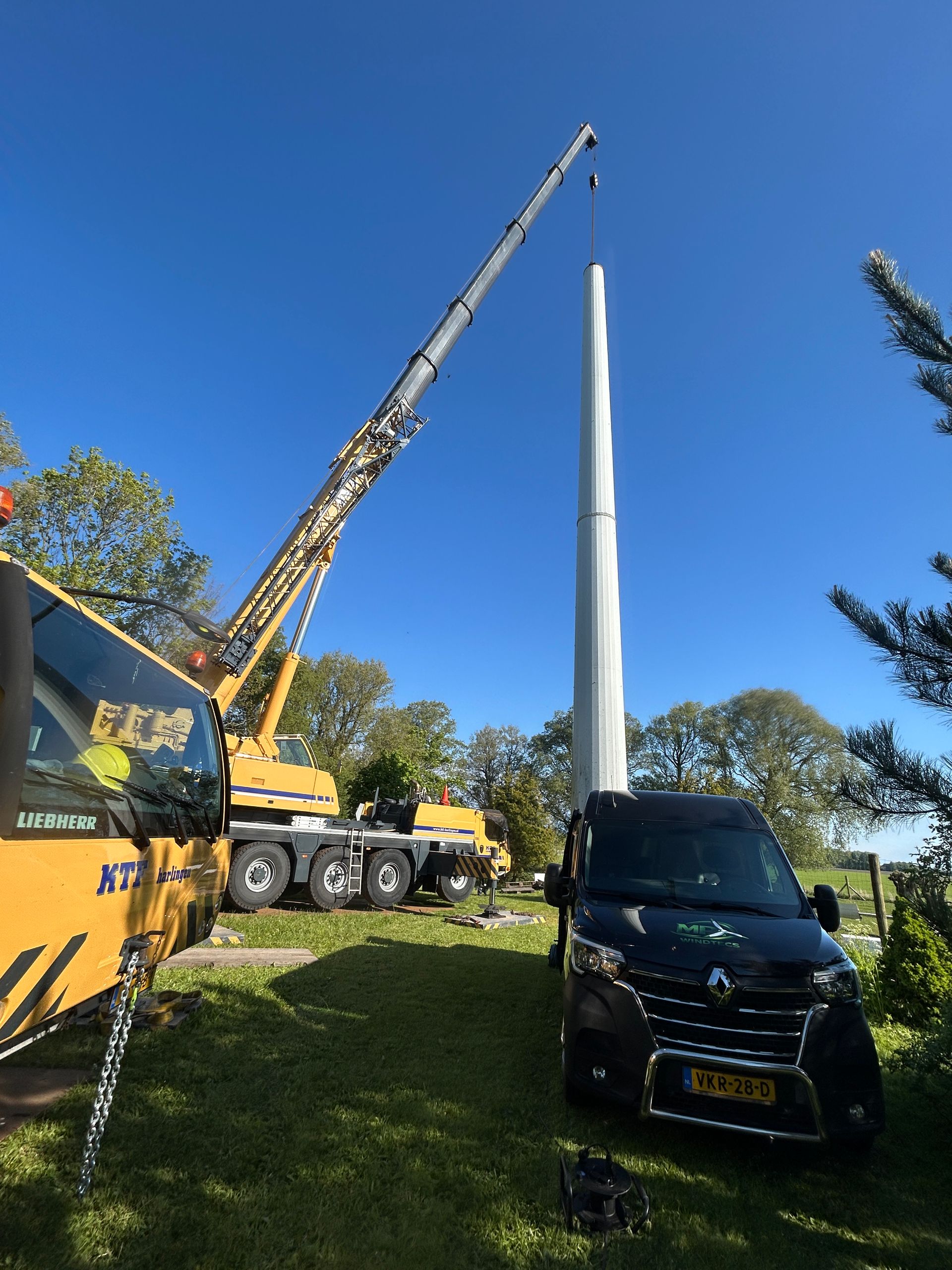 Crane lifting a tall white tower beside a black van on a sunny grassy site