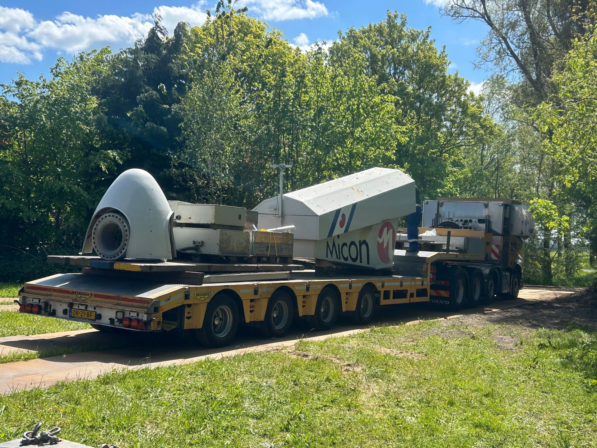 Oversized industrial machine on a lowboy trailer parked on a grassy roadside under trees and blue sky