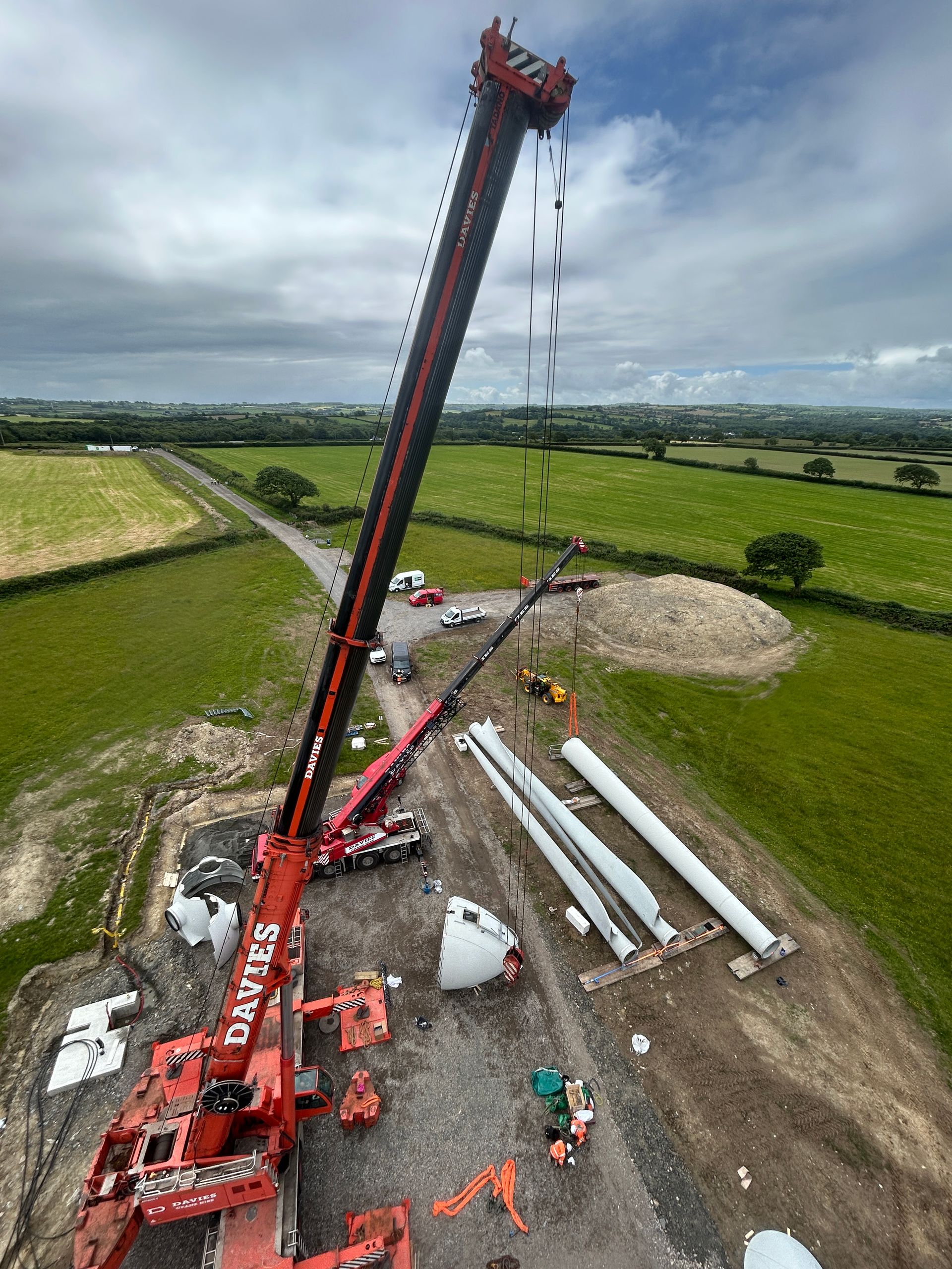 Red crane lifting a long dark beam over stacked white pipes in a rural construction site