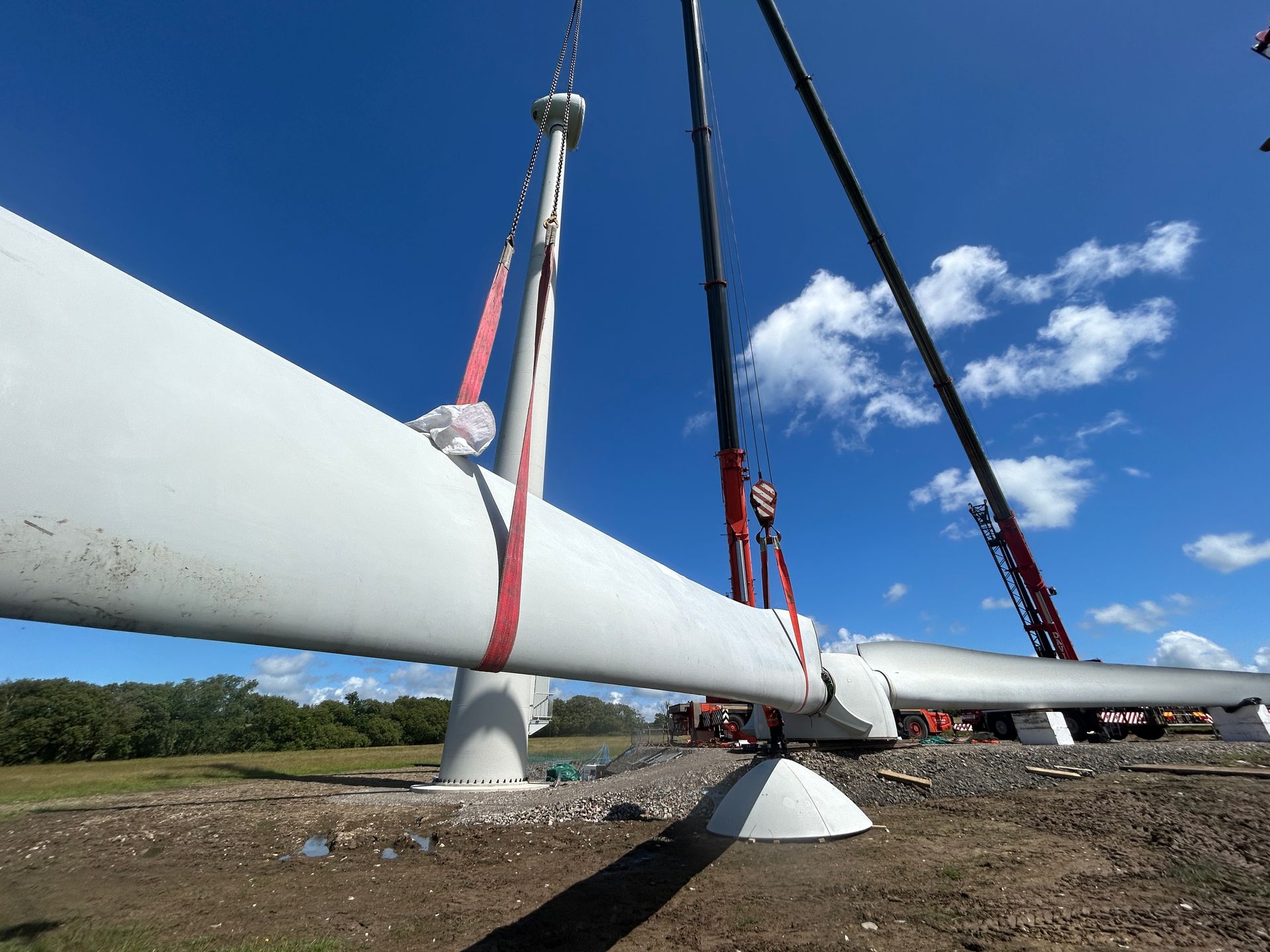 Wind turbine blade being lifted by cranes at a construction site under a blue sky.