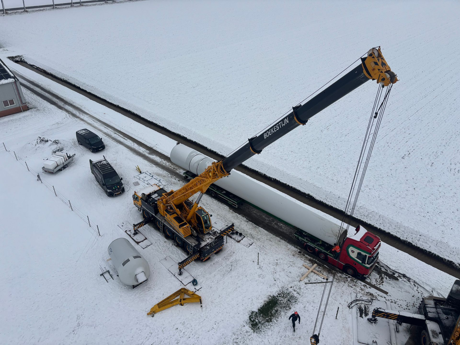 Aerial view of a crane lifting a black pipeline beside a red truck on a snowy construction site.