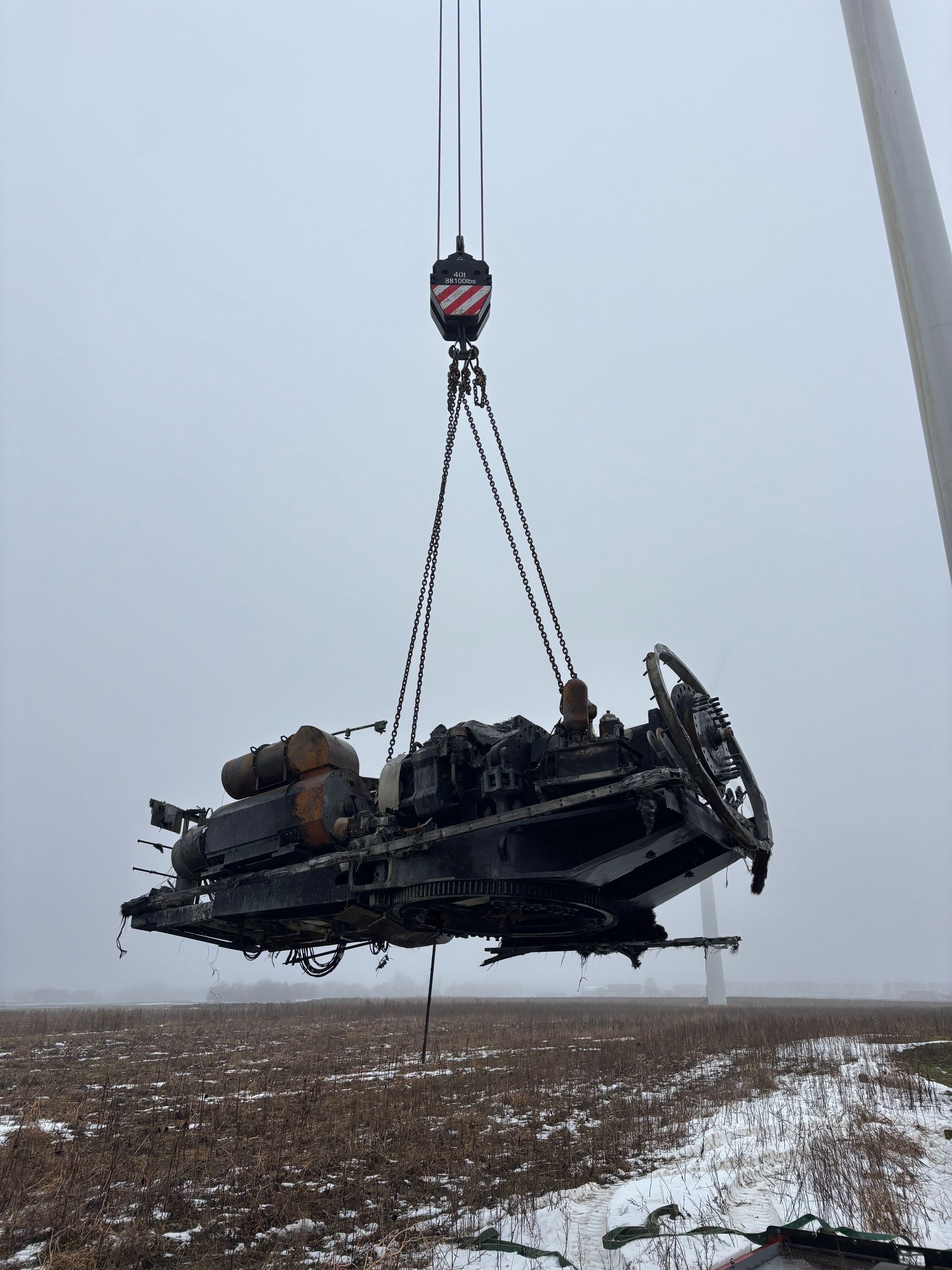 Large black vehicle suspended by crane over a snowy, muddy field in foggy conditions