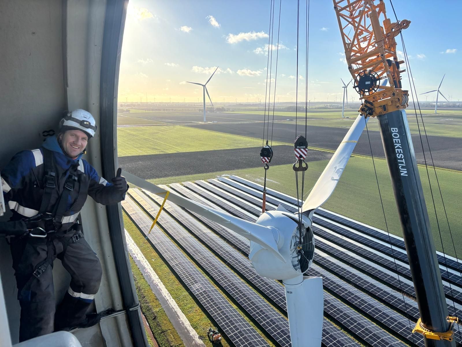 Worker in helmet on wind turbine platform above a field at sunrise, with orange crane and cables nearby