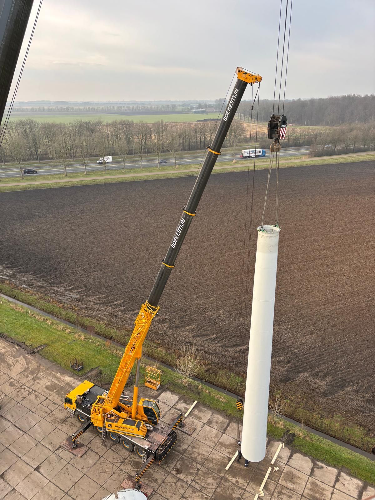 Orange-and-black utility post driver leaning beside a white pole on a roadside, with an open field in the background