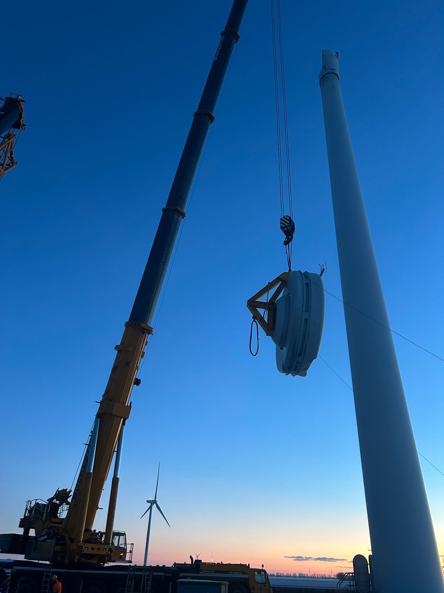 Crane lifting a wrapped load beside a tall wind turbine at sunset, with a clear blue sky.