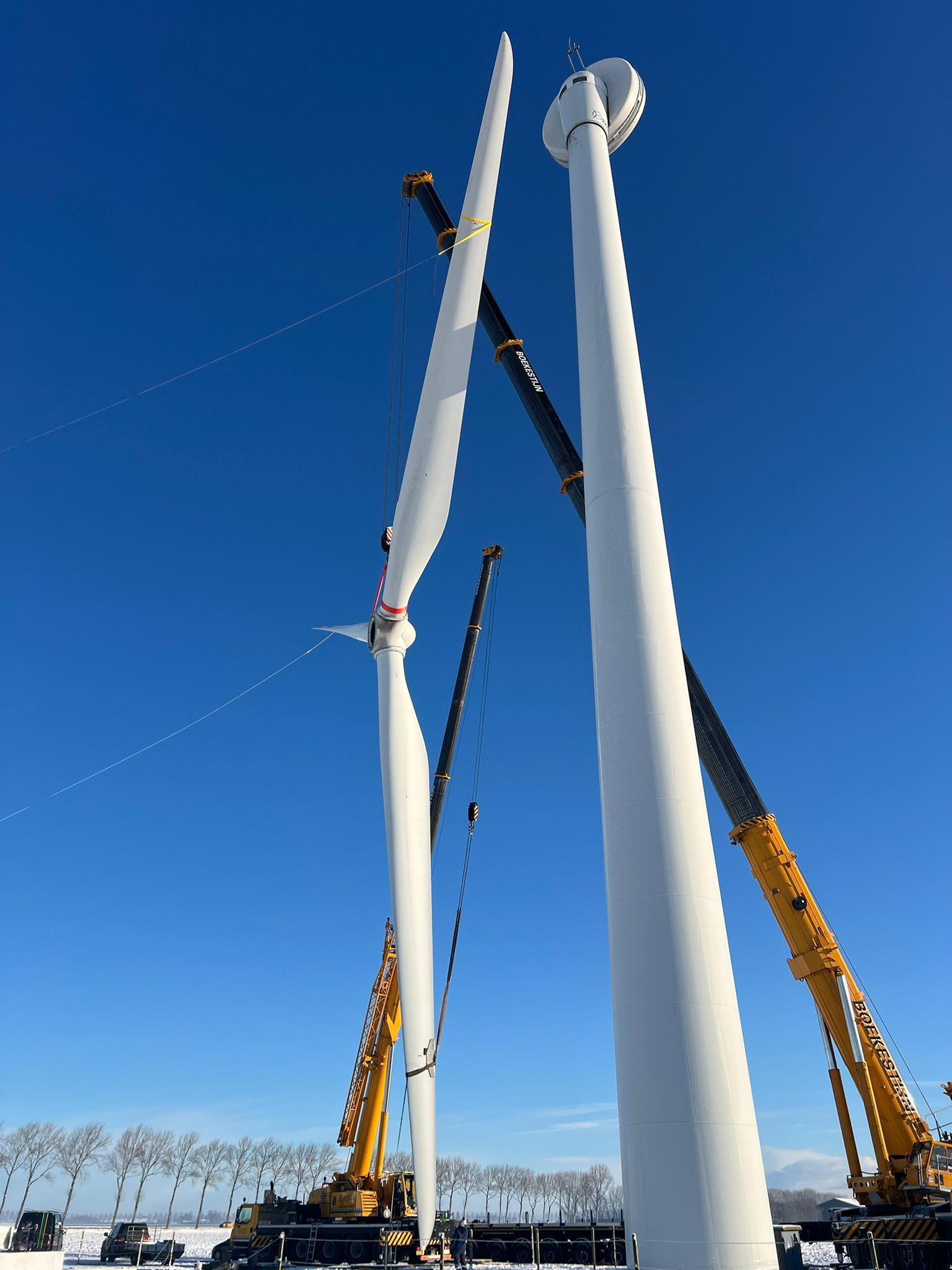 Two large white wind turbine towers being installed by cranes under a clear blue sky