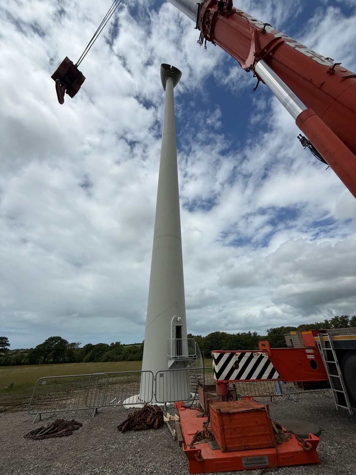 Crane lifting a tall white wind turbine tower section at a construction site under cloudy sky