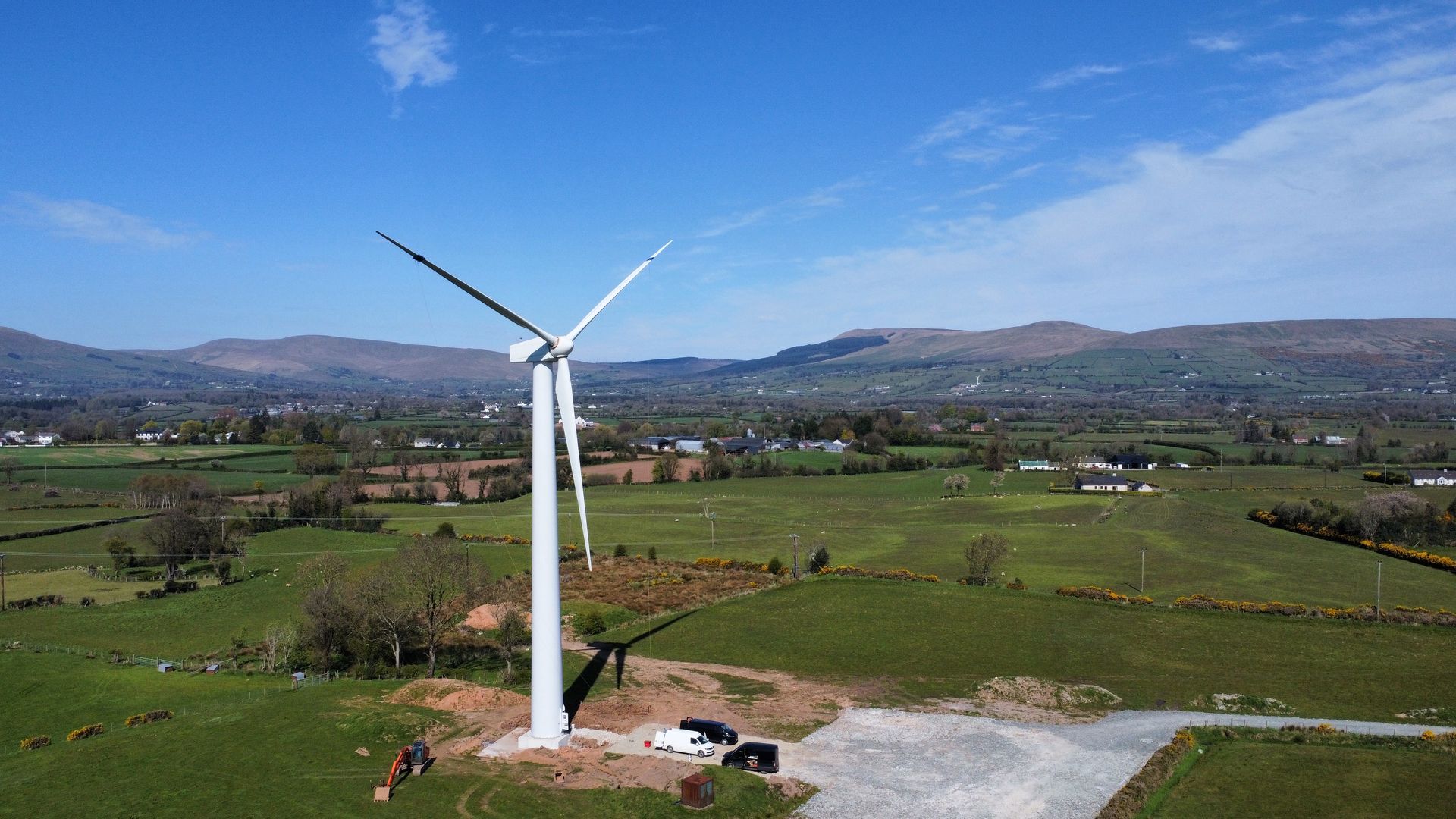 White wind turbine in a green rural field with mountains under a clear blue sky