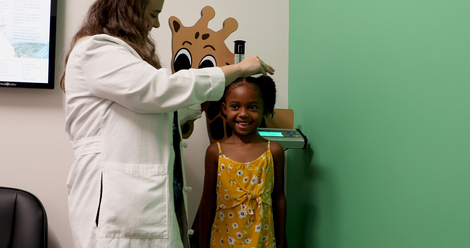 A medical professional measuring a child's height against a giraffe-themed scale in a clinic.