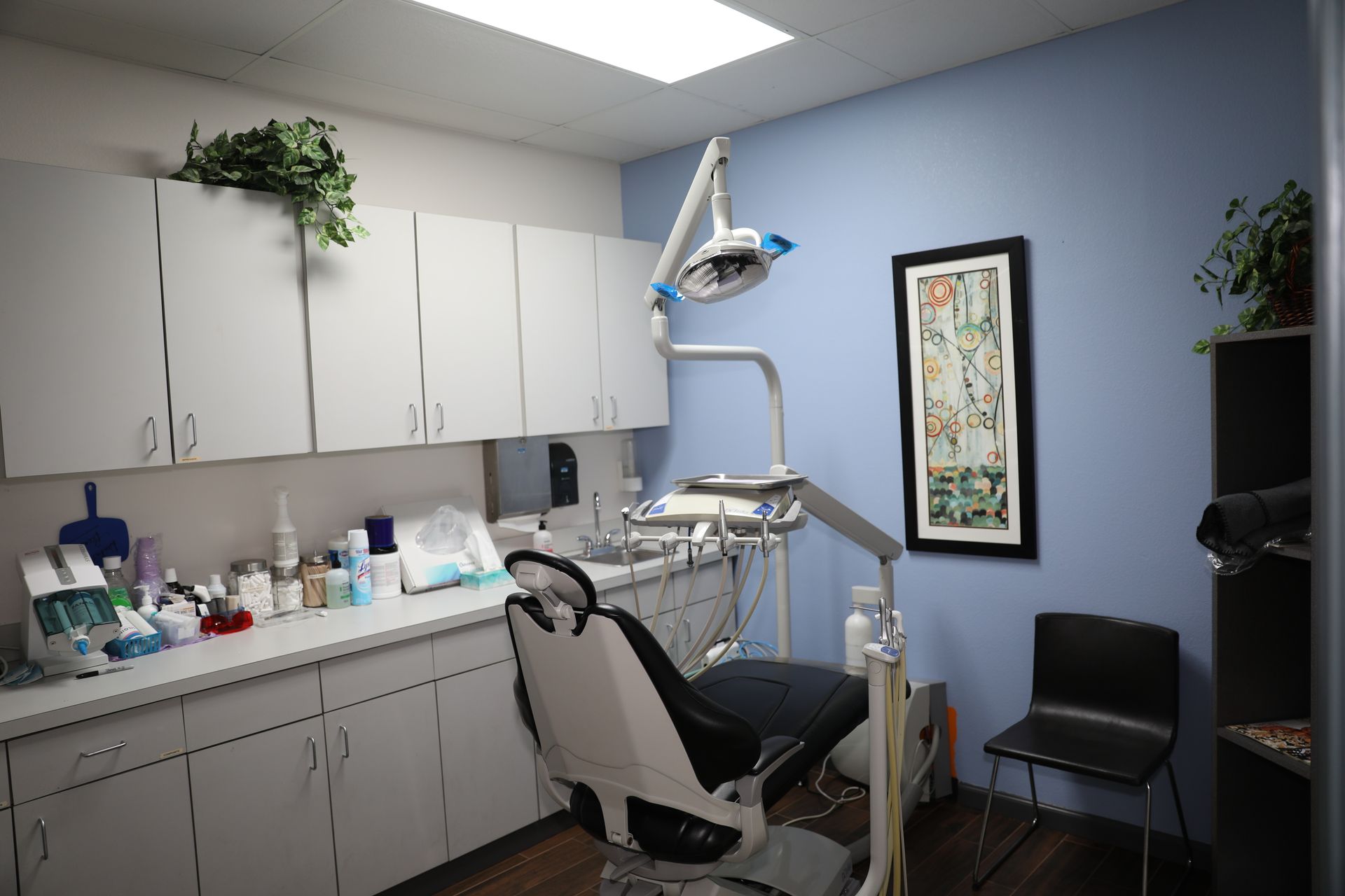 A dental examination room with a patient chair, dental equipment, white cabinets, and blue walls.
