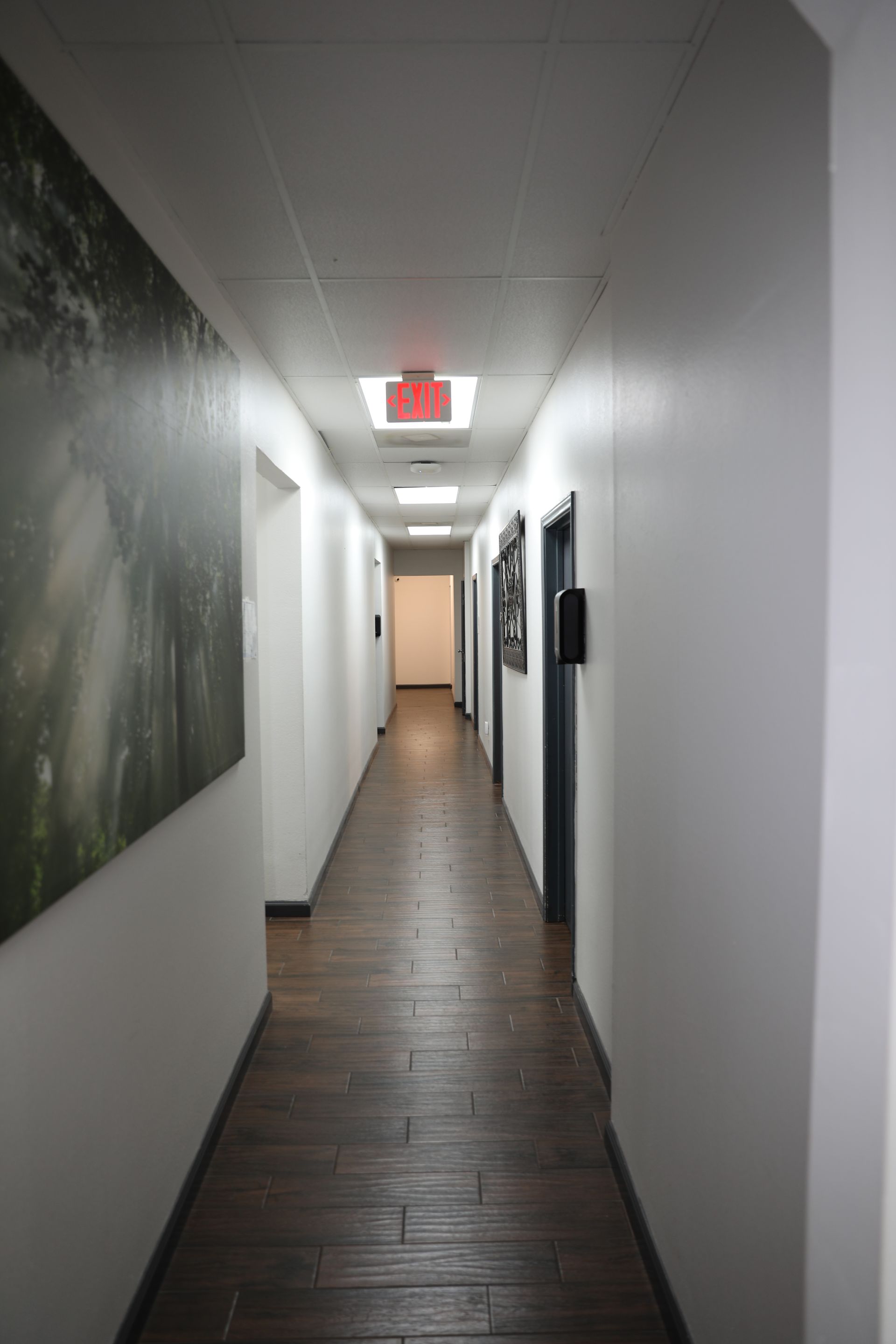 A long, narrow indoor hallway with dark wood-look tile flooring, white walls, and a red exit sign on the ceiling.