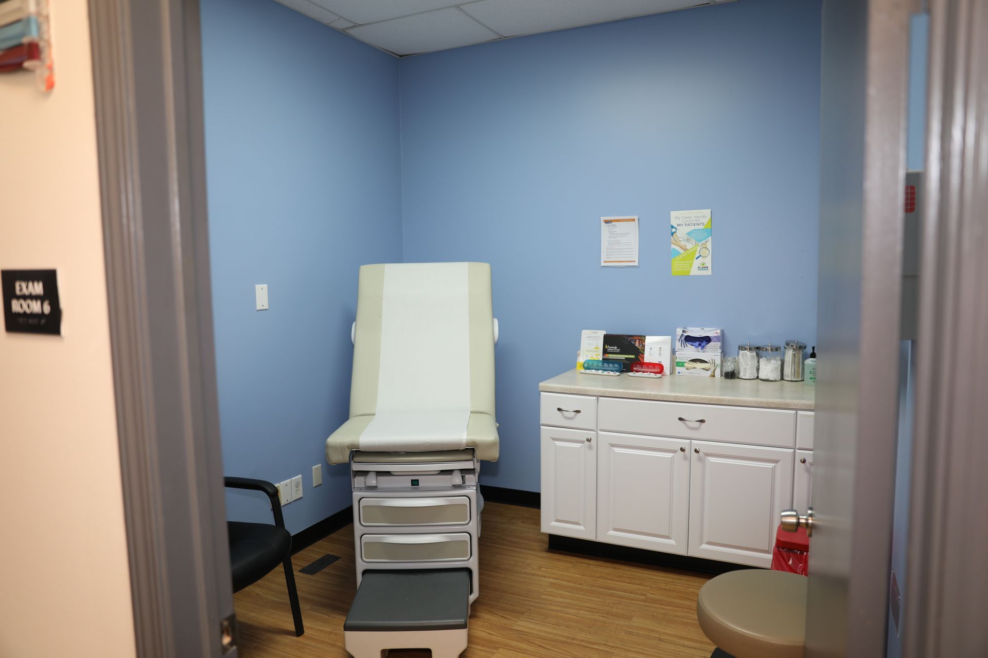 A medical exam room with blue walls, a white examination table, a black chair, and white storage cabinets.