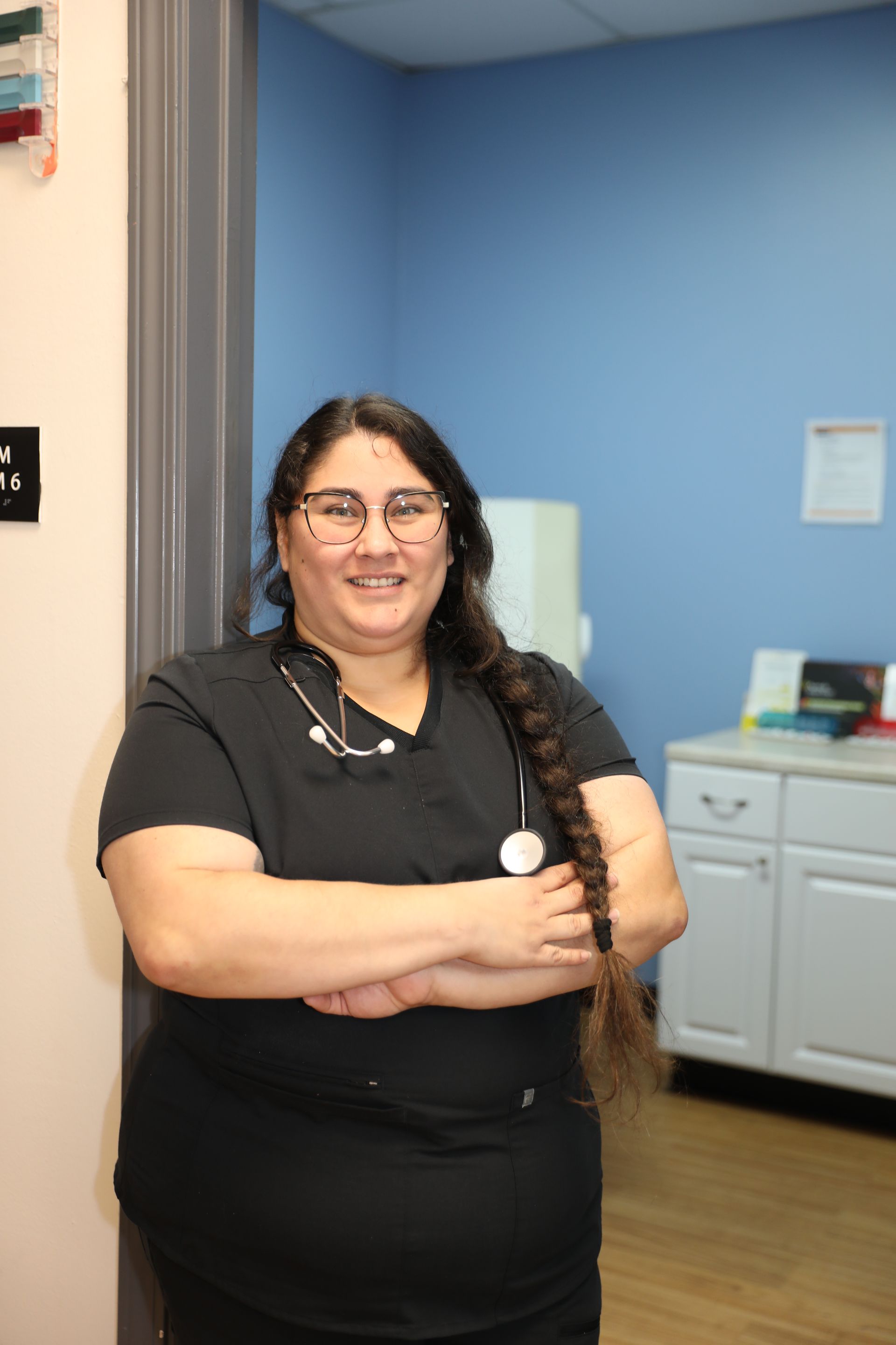 A person wearing black medical scrubs and glasses with a stethoscope around their neck, smiling in a clinical office.