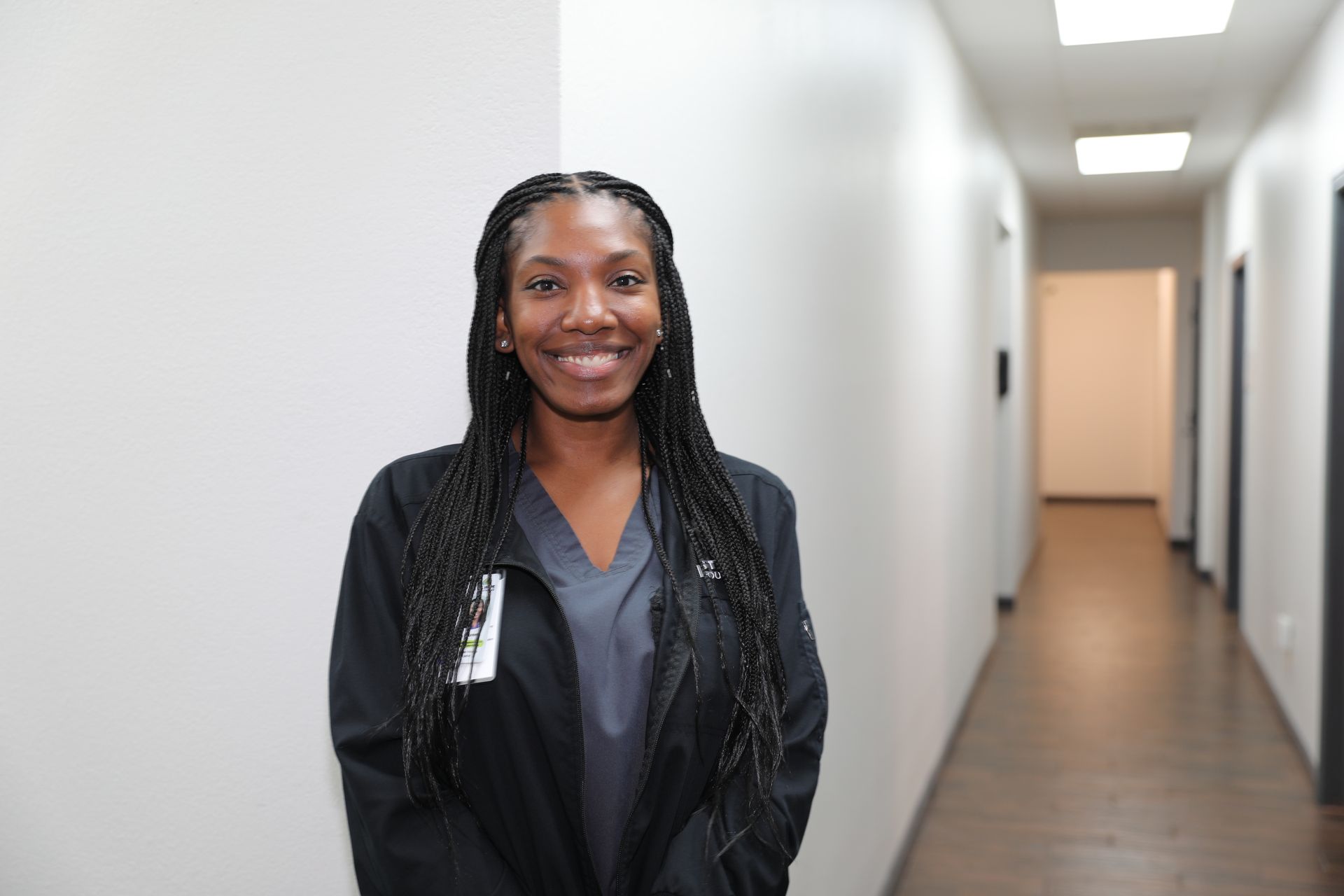 A woman wearing scrubs and a black jacket smiles while standing in a brightly lit office hallway.