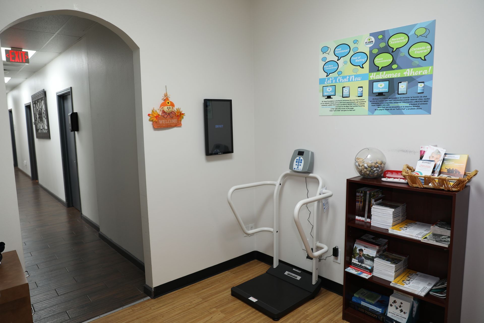 A medical scale stands in a hallway office next to a bookshelf, with an informational poster on the wall.