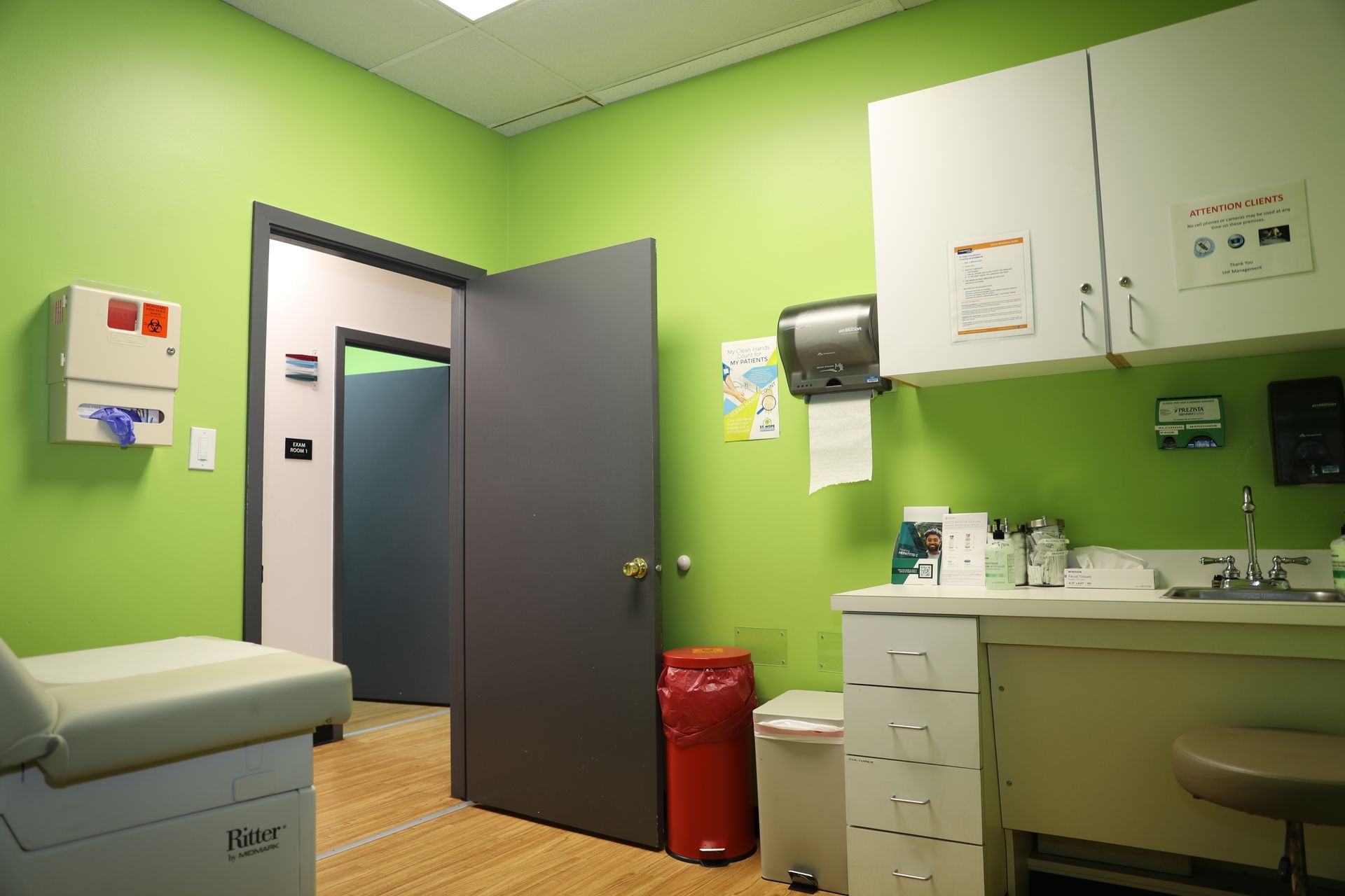 A lime-green medical exam room with a grey door, exam table, sink, cabinets, and waste bins.