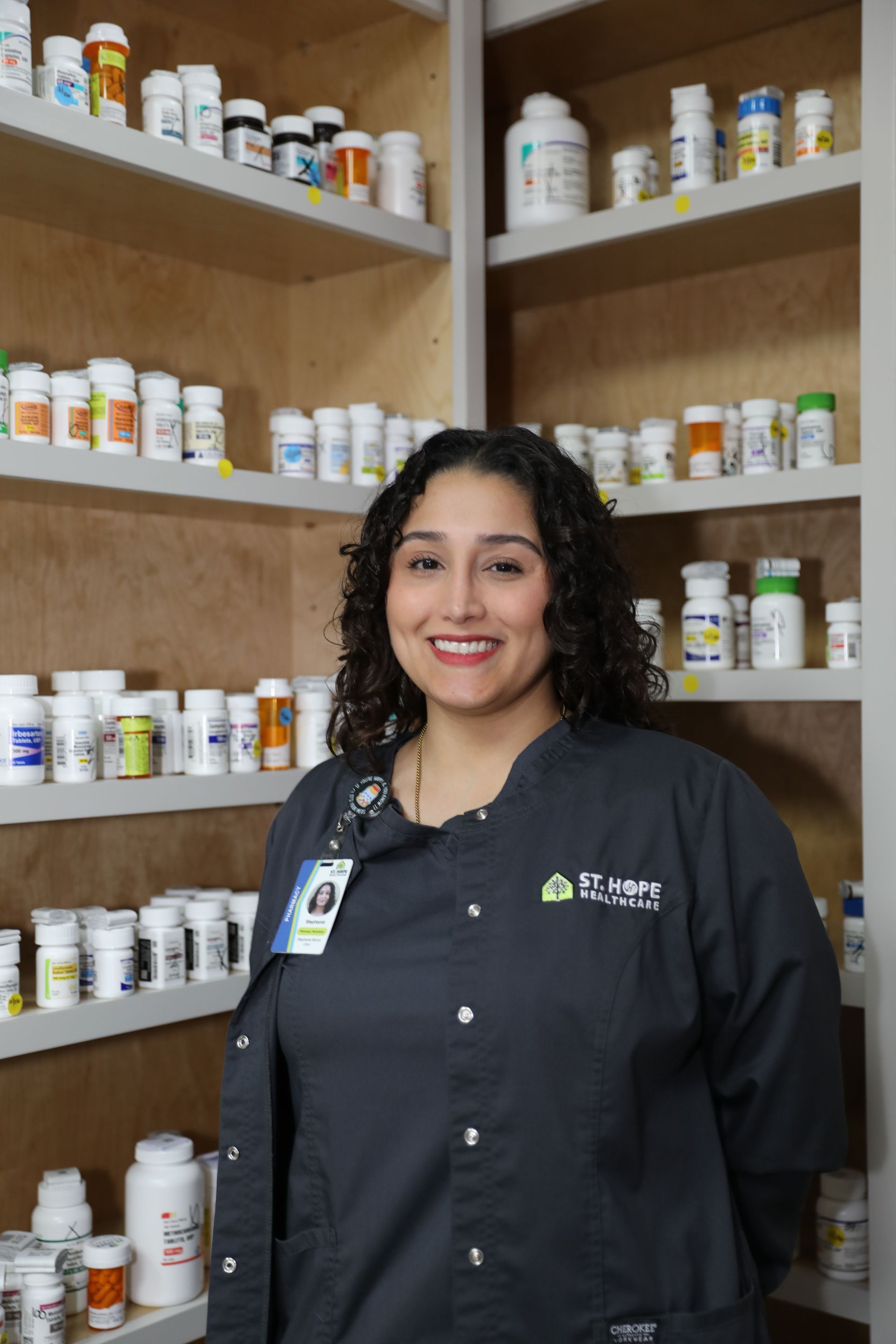 A person wearing a gray pharmacy work shirt stands smiling in front of shelves stocked with various prescription bottles.