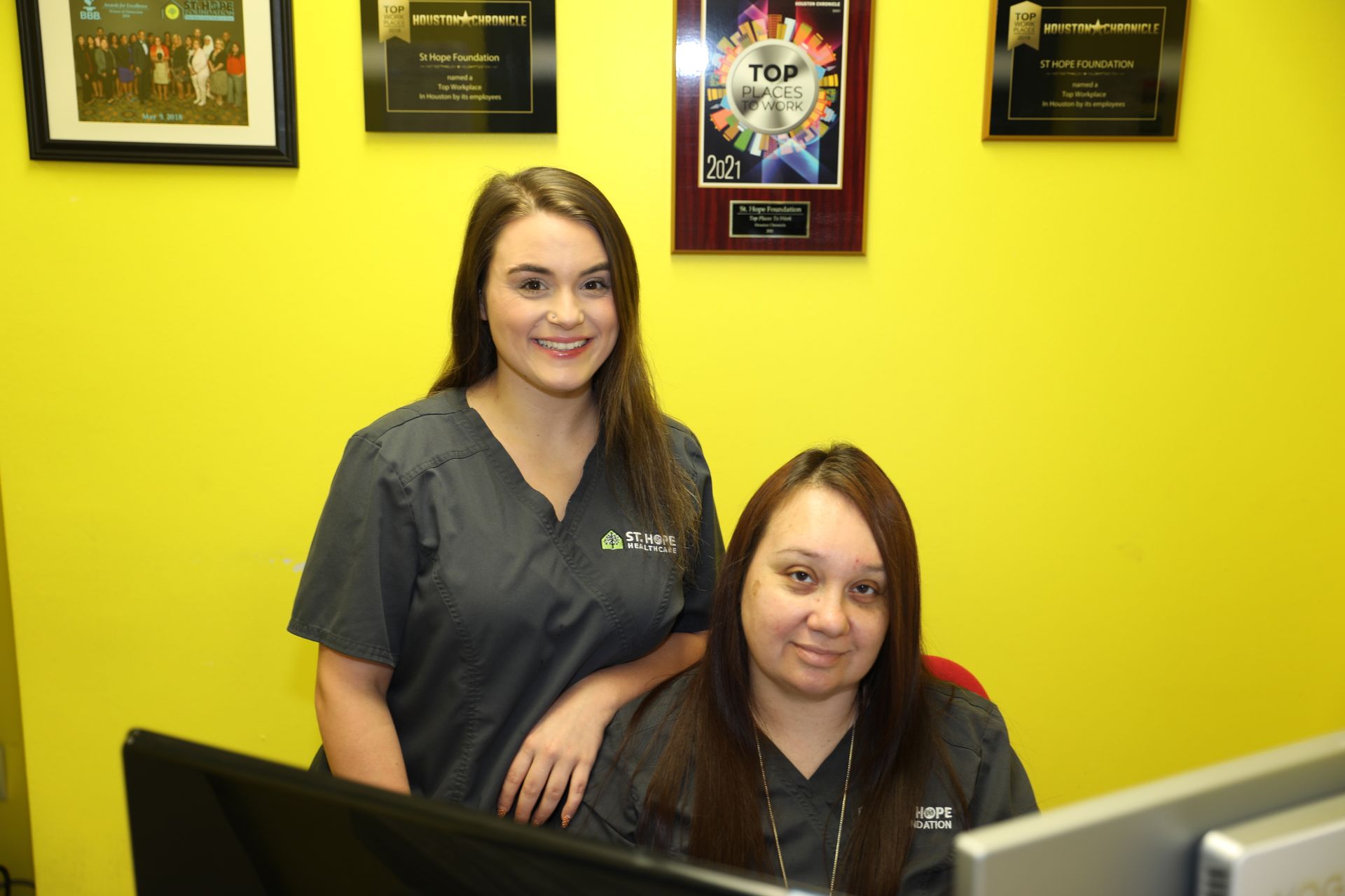 Two staff members in dark scrubs pose behind a desk against a yellow wall adorned with framed awards.
