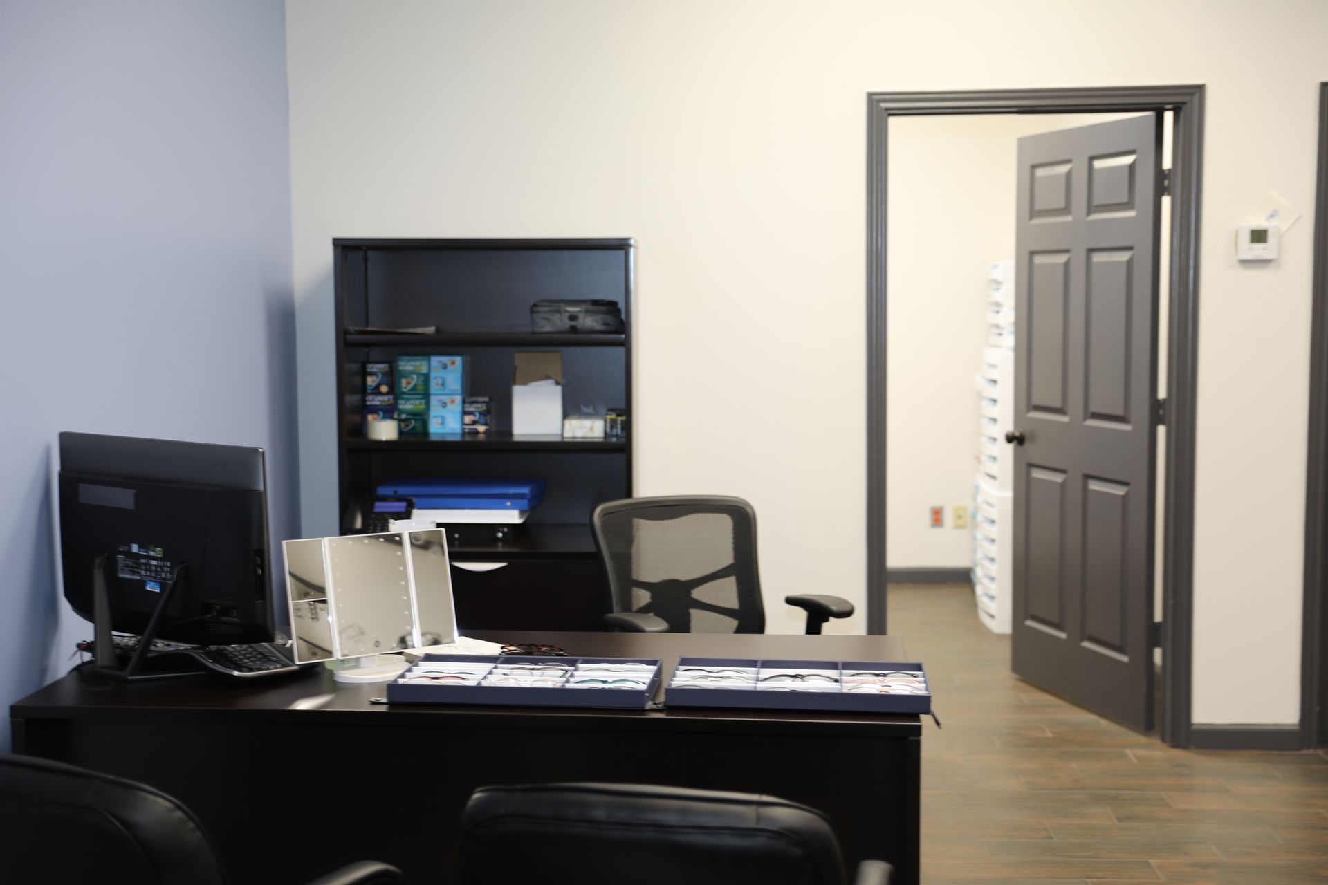 A modern office with a dark desk, computer, shelves, a chair, and an open doorway leading to an adjacent room.