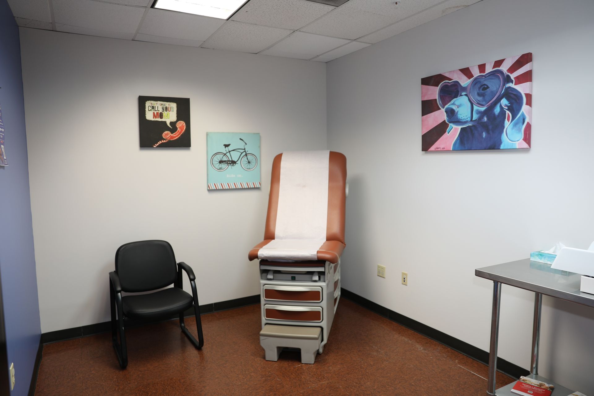 A medical exam room featuring a brown exam table, a black chair, and three decorative wall prints.