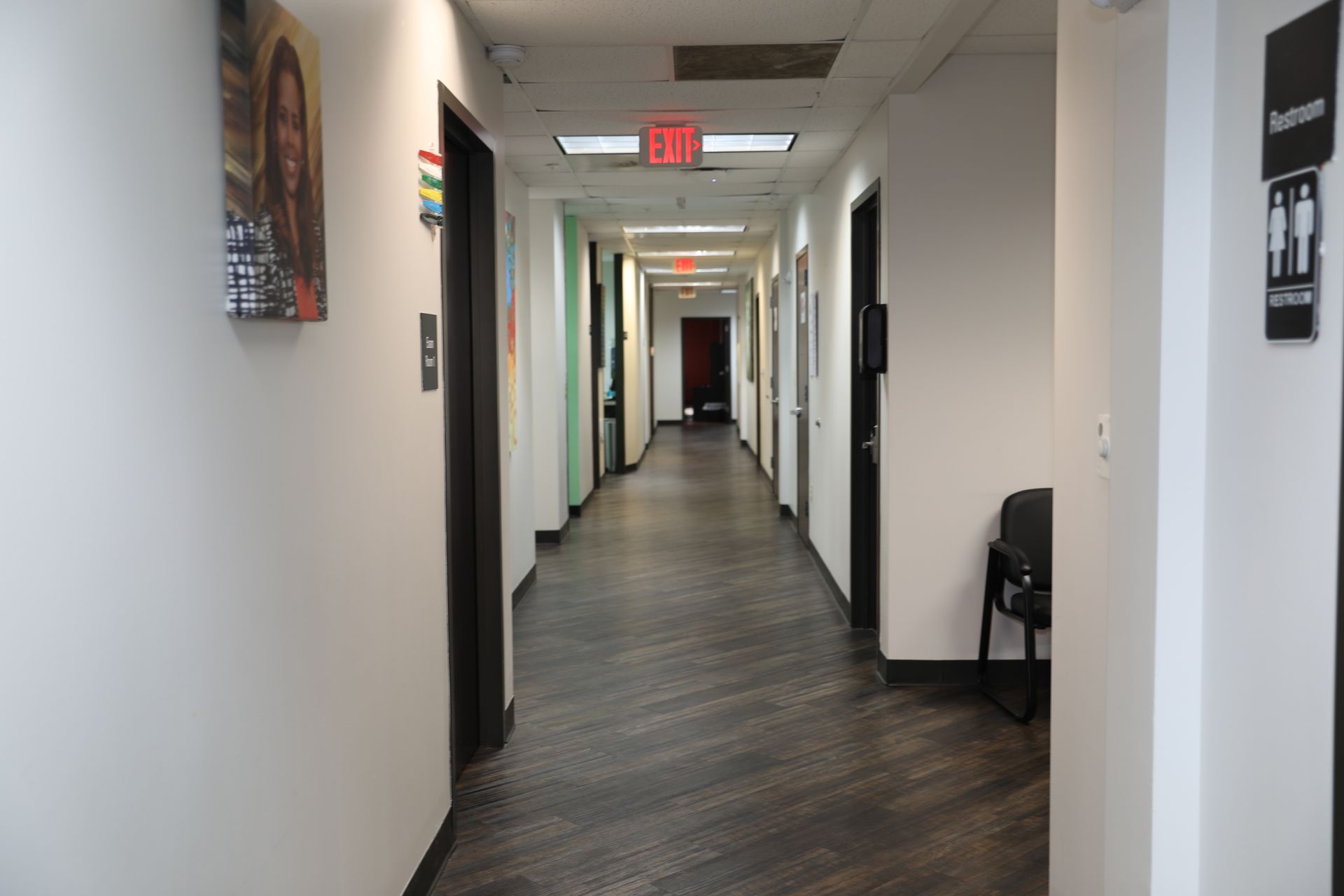 An indoor office hallway with white walls, dark wood-look flooring, multiple doorways, and an overhead exit sign.