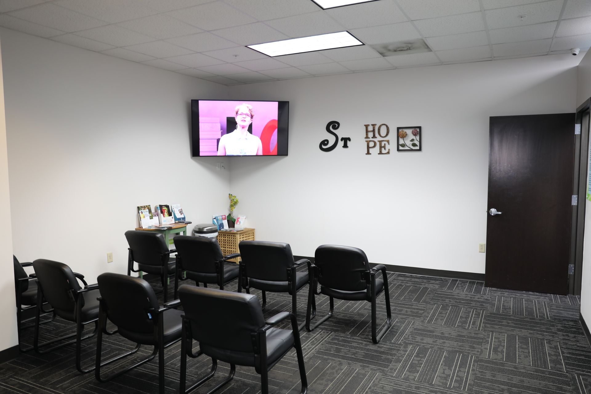 A small office waiting room with black chairs arranged in rows, a wall-mounted TV, and a sign reading 