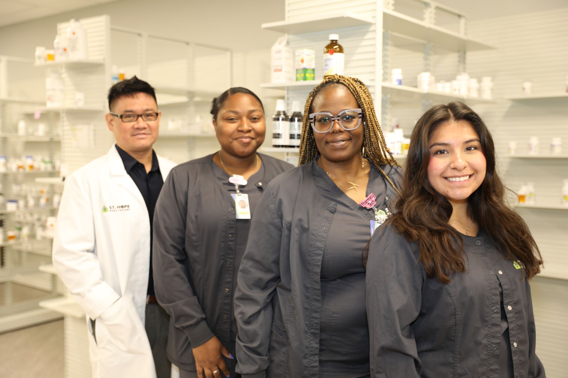 Four pharmacy staff in professional attire, including a lab coat and scrubs, posing together in front of medicine shelves.