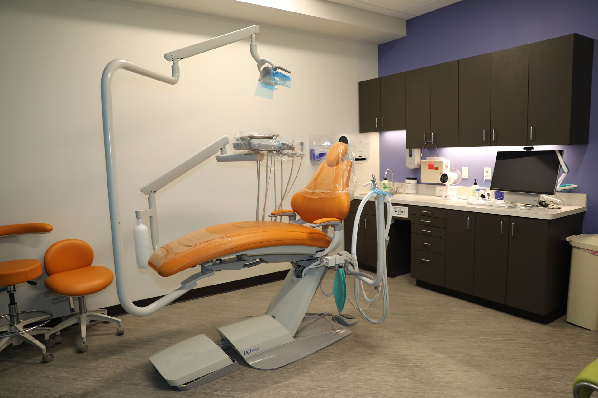 A dental operatory room featuring an orange dental chair, a diagnostic light, dark cabinetry, and two rolling stools.