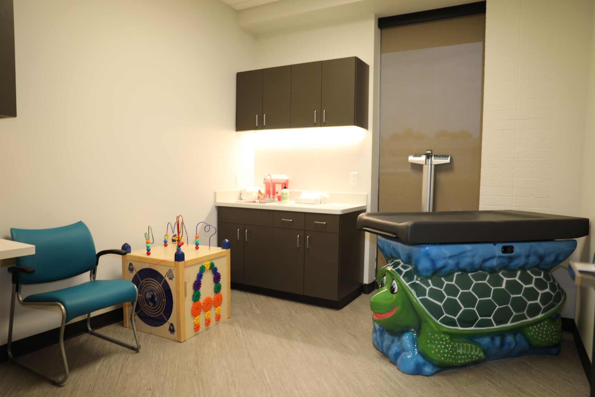 A pediatric examination room featuring a turtle-shaped exam table, a wooden activity cube, and a blue chair.