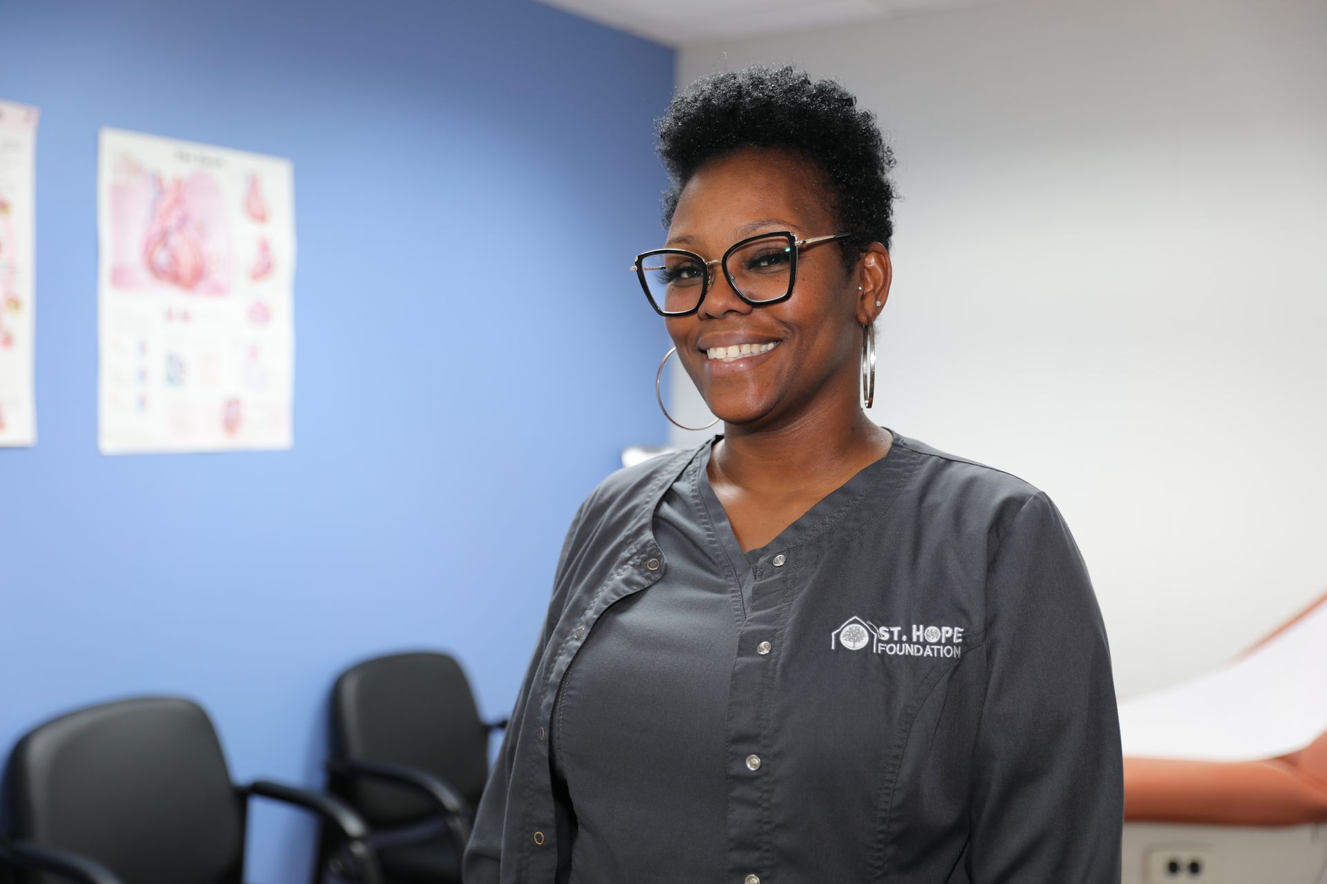 A person with short hair and glasses wearing a gray medical scrub top, smiling in a clinical office setting.