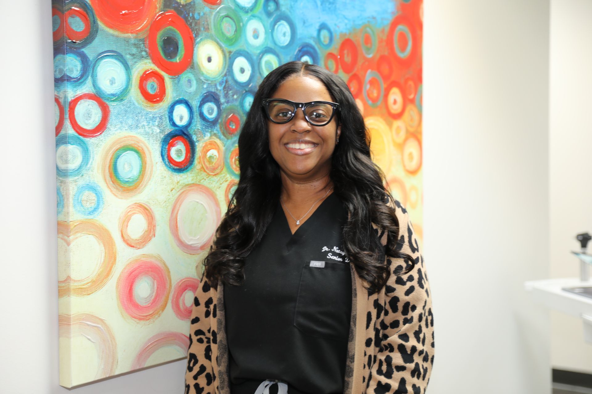 A professional smiling, wearing glasses and a dark scrub top with a leopard print cardigan, standing before abstract art.