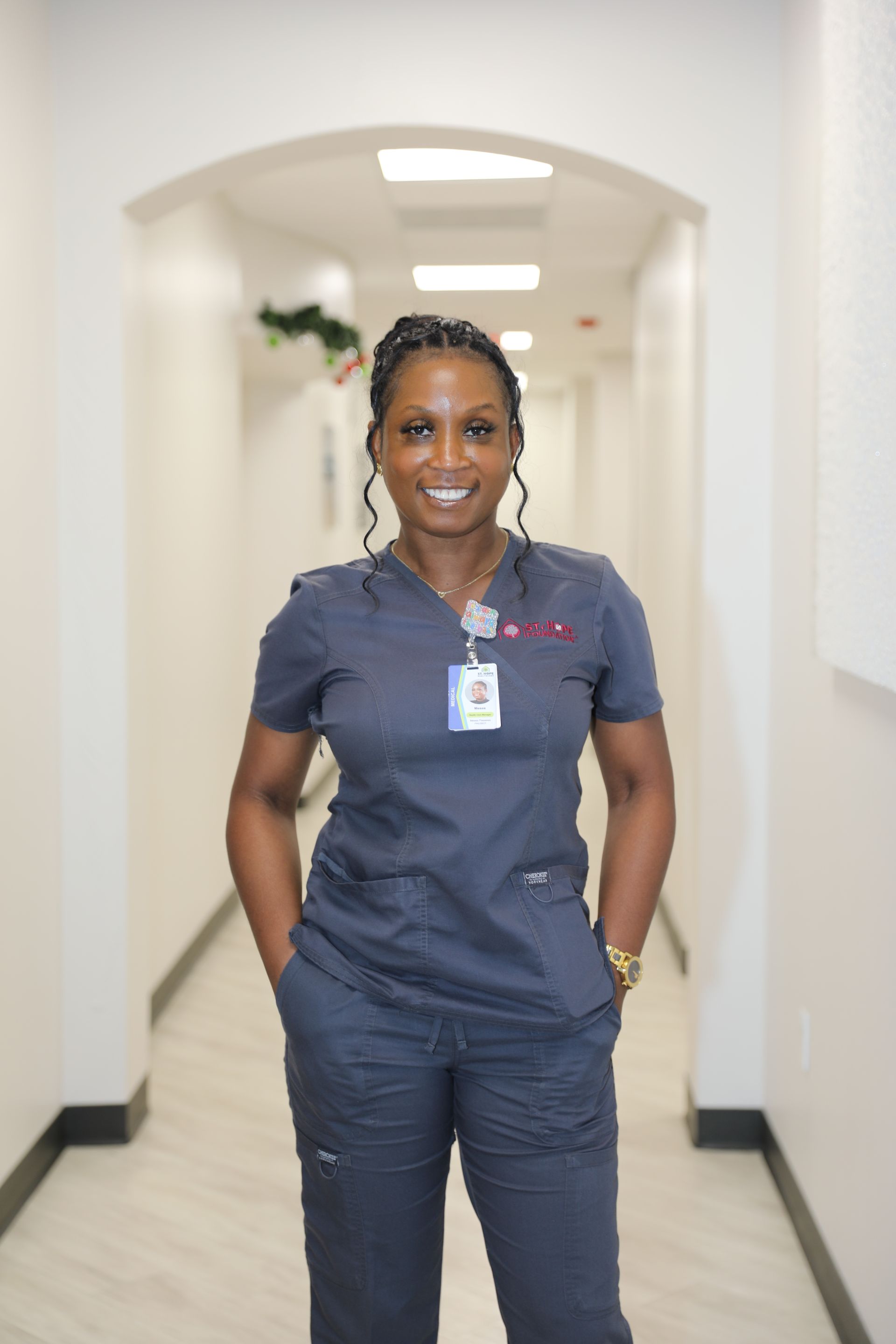 A person wearing grey medical scrubs and a name tag, smiling while standing in a brightly lit hallway.