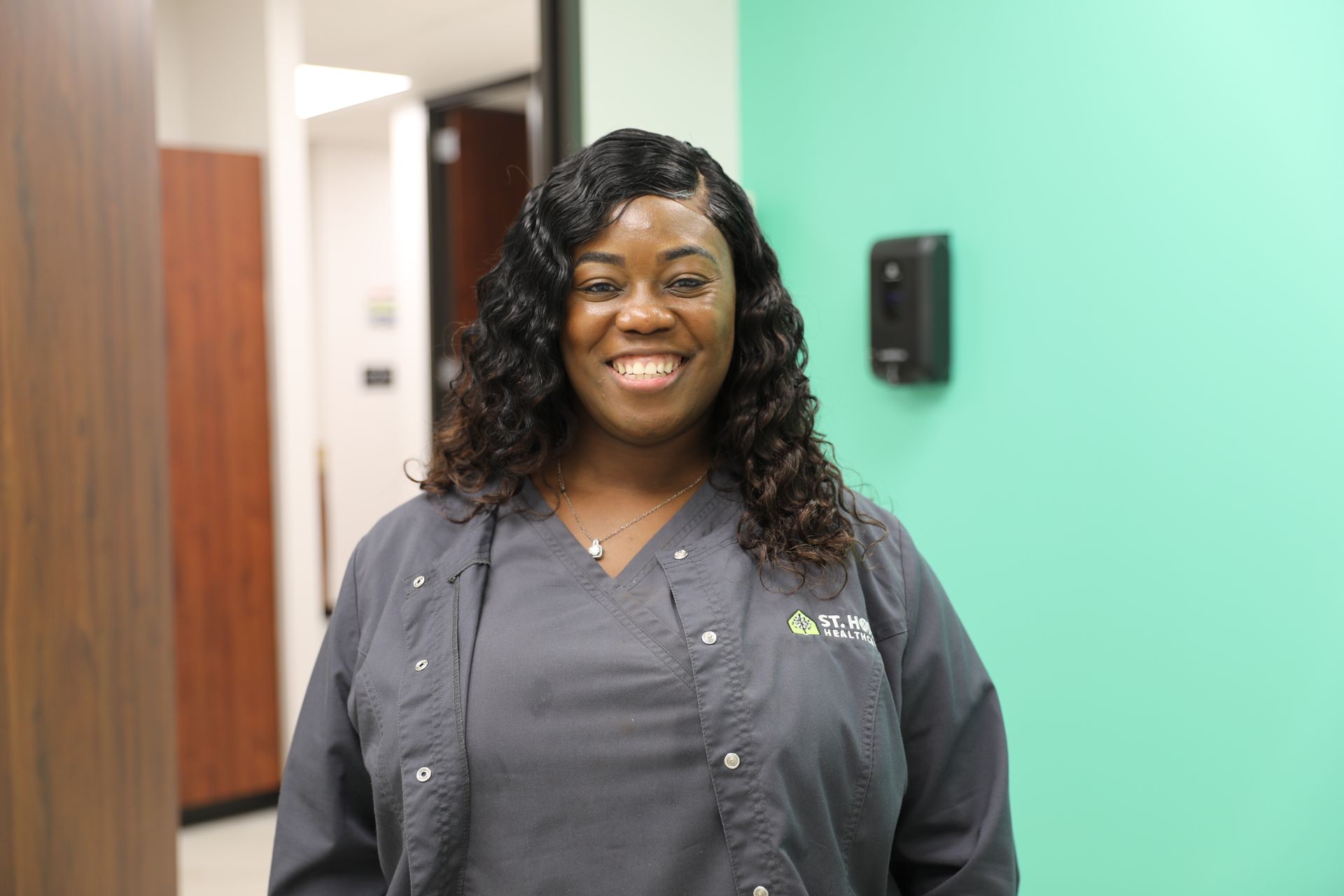 A smiling professional wearing a grey scrub top stands in an office hallway with a light green wall and dispenser.