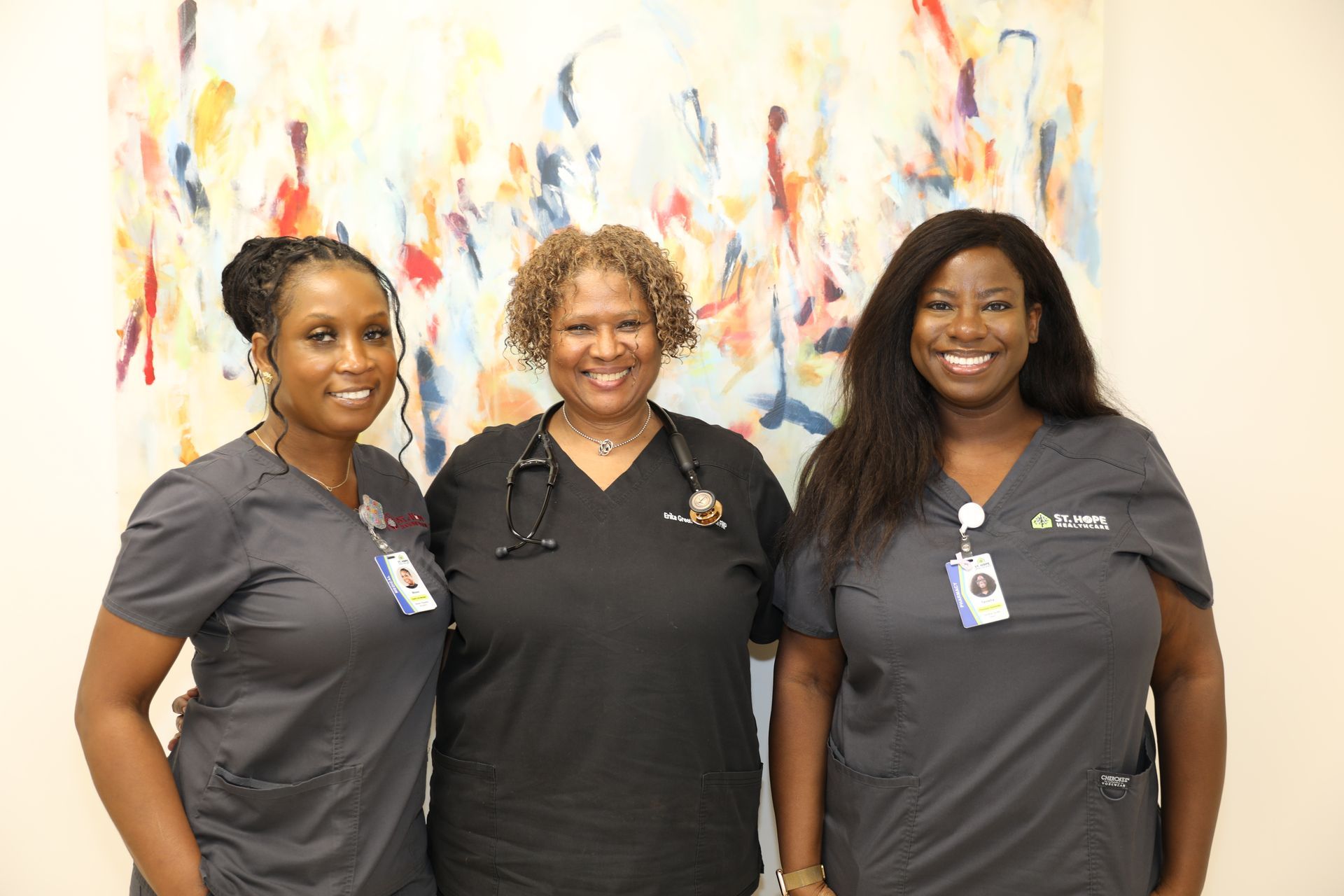 Three smiling medical professionals in gray scrubs stand side-by-side in front of an abstract, colorful wall mural.