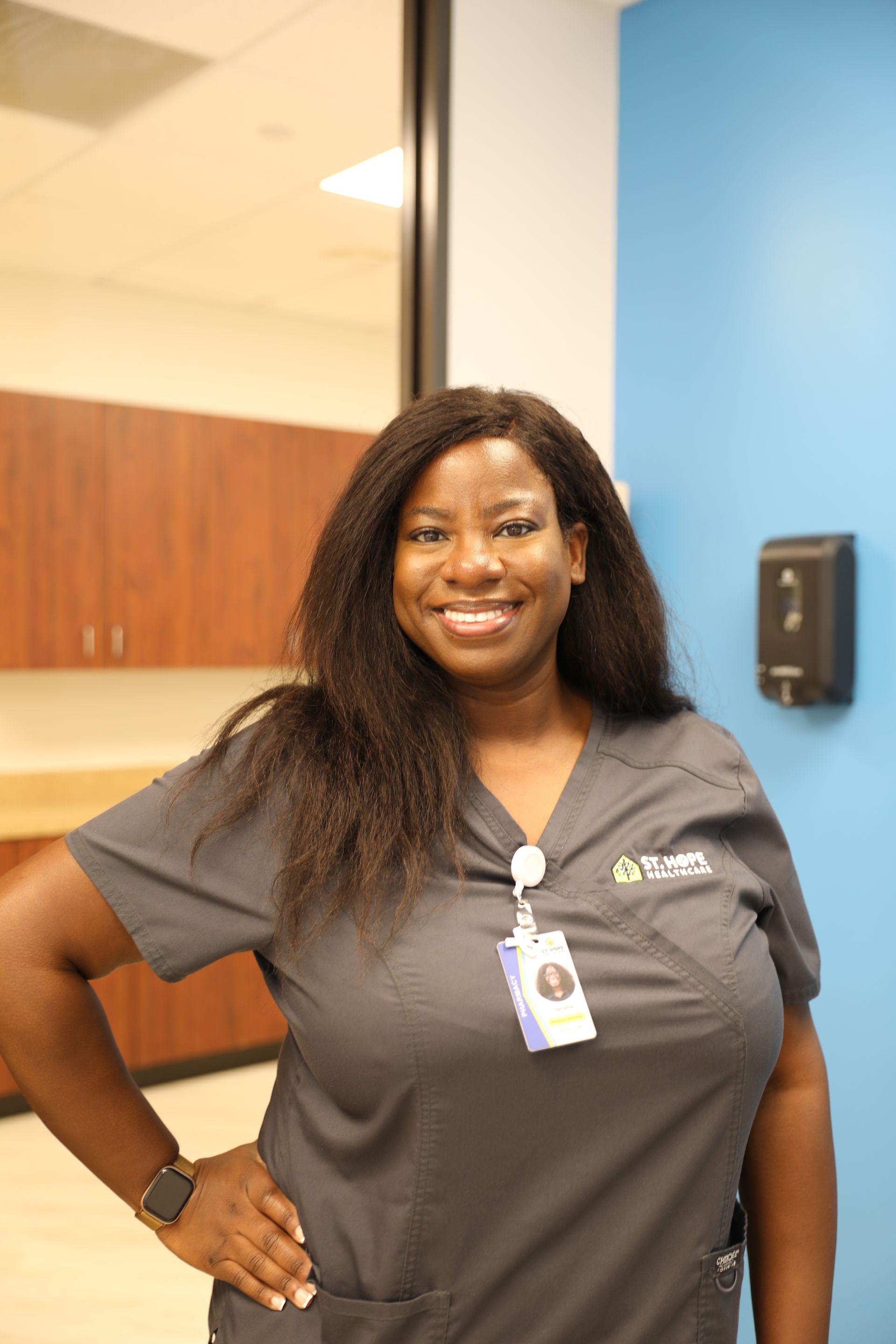 A smiling professional in a dark grey medical scrub top with an ID badge stands against a blue wall in an office.
