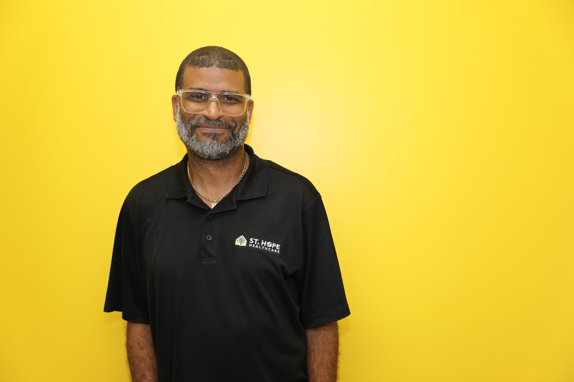 A person with a beard and glasses smiles, wearing a black polo shirt against a bright yellow background.