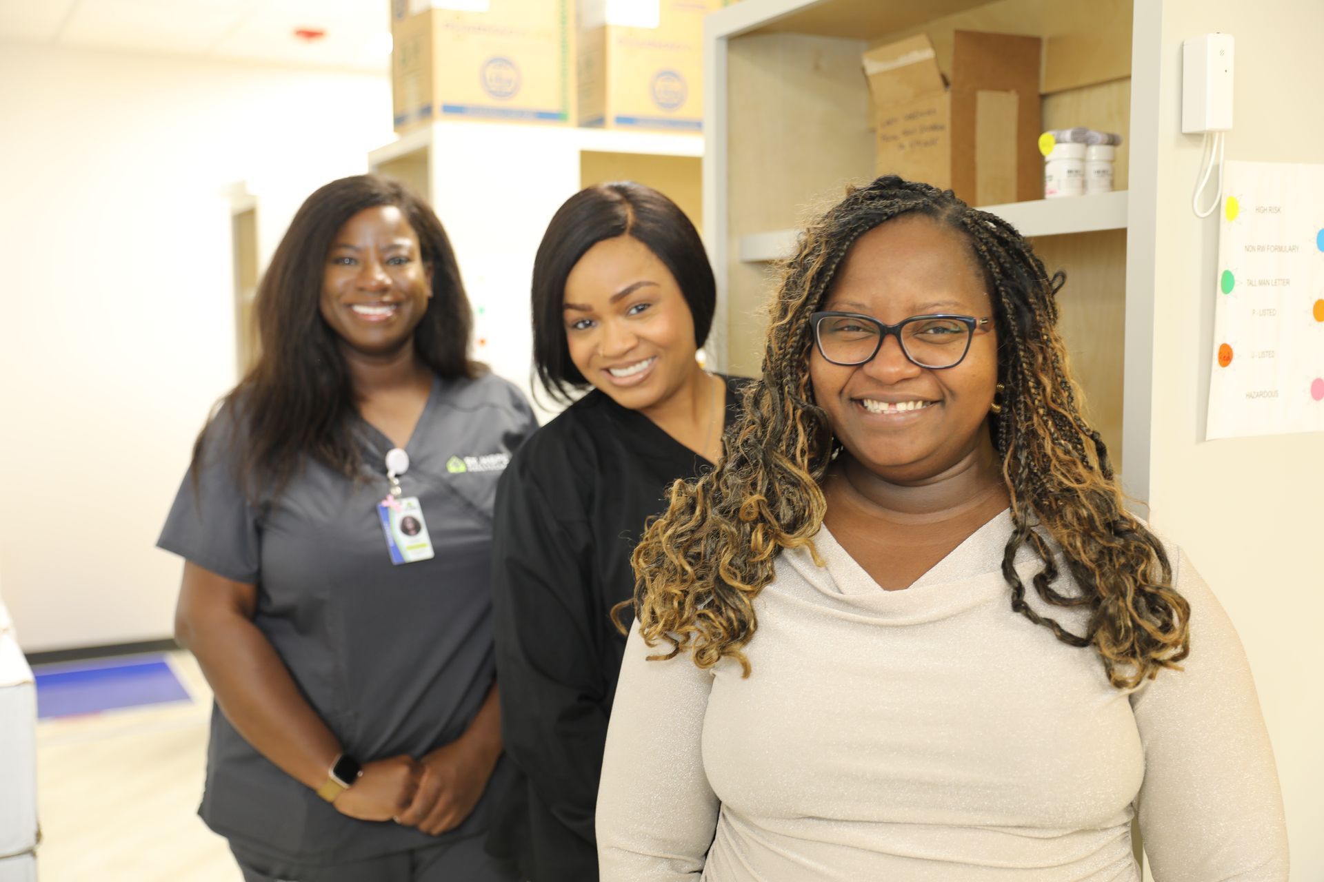 Three people wearing professional attire smile while posing in an office or clinic setting with shelving in the background.