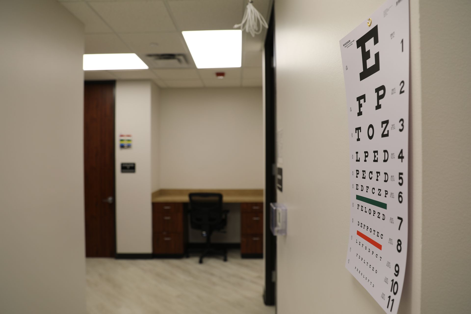 A snellen eye chart mounted on a white wall in the foreground, with an out-of-focus office space in the background.