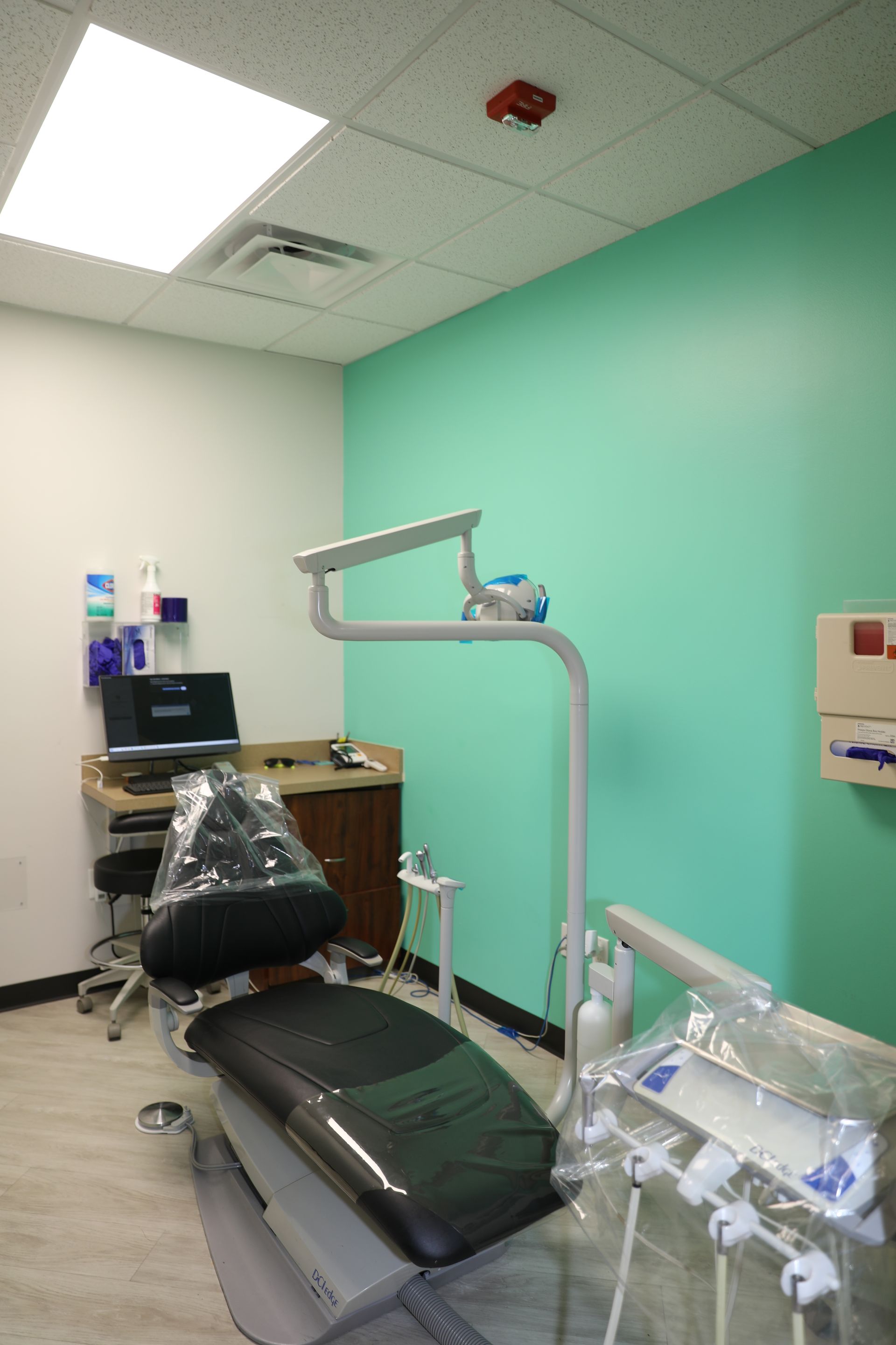 A dental examination room with a black reclining chair, adjustable overhead light, and medical equipment on a counter.