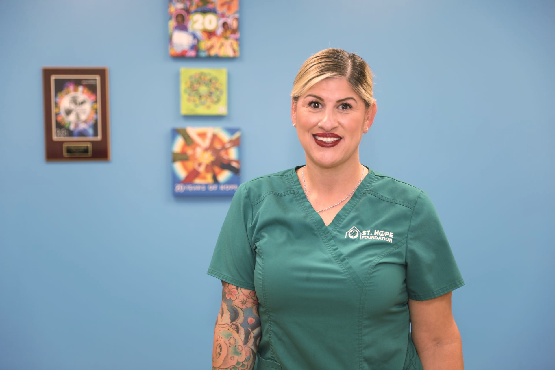 A smiling person in a green scrub top with arm tattoos standing in front of a blue wall with three framed art pieces.