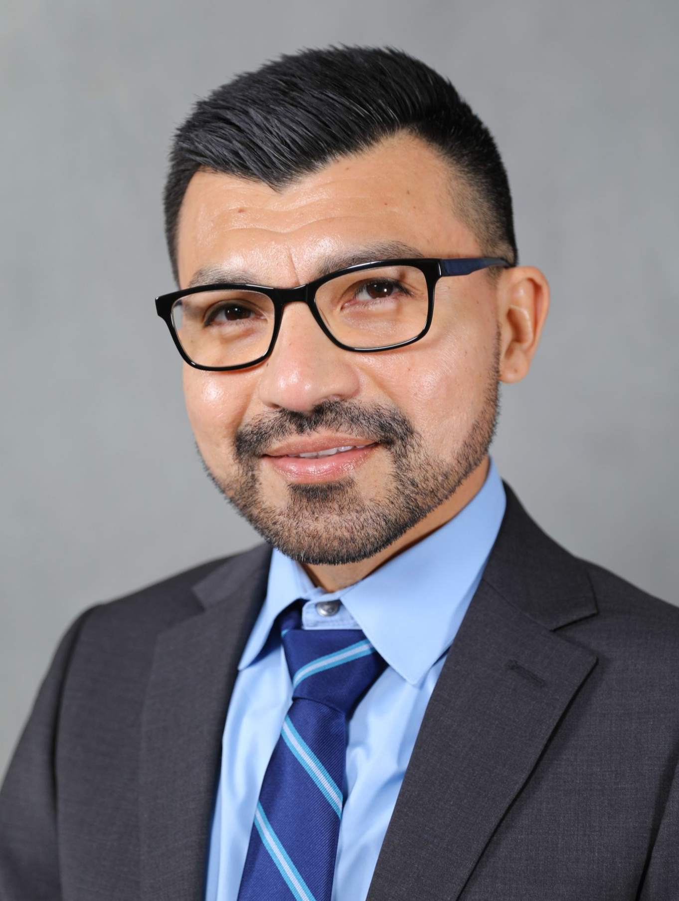 A professional portrait of a person with a beard and glasses, wearing a dark suit, light blue shirt, and blue tie.