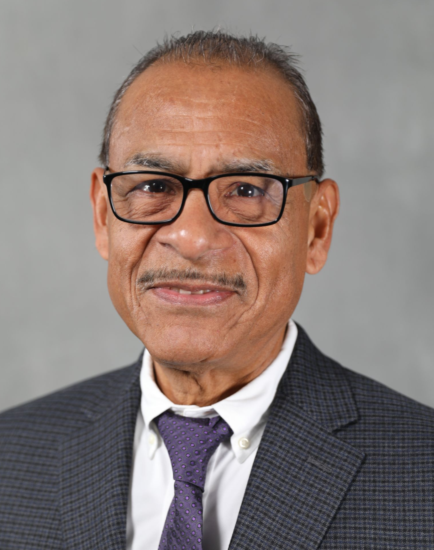 A man wearing glasses, a white collared shirt, and a dark patterned tie and blazer against a plain gray background.