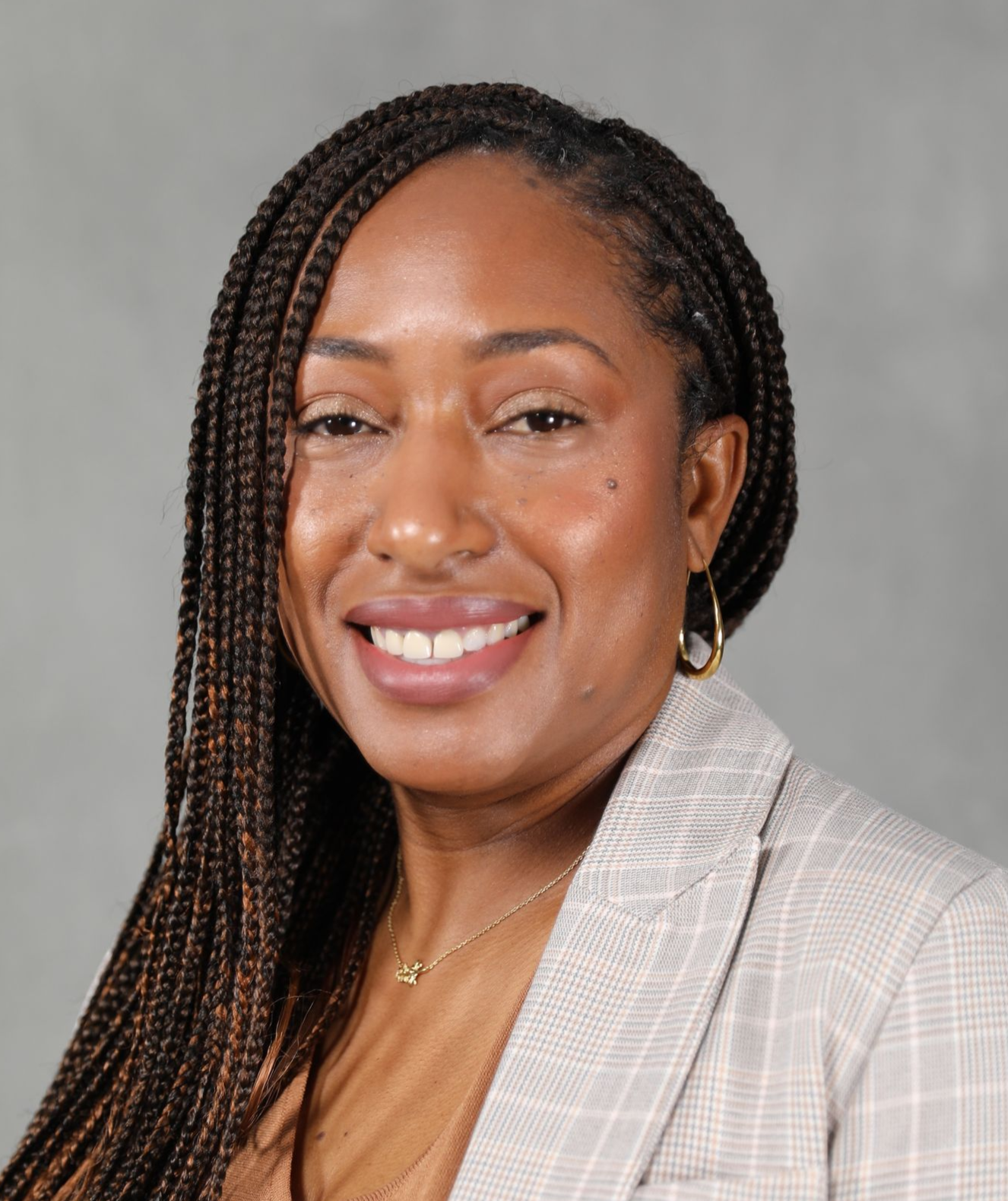 A professional headshot of a person with long braided hair, smiling, wearing a light-colored blazer against a grey backdrop.