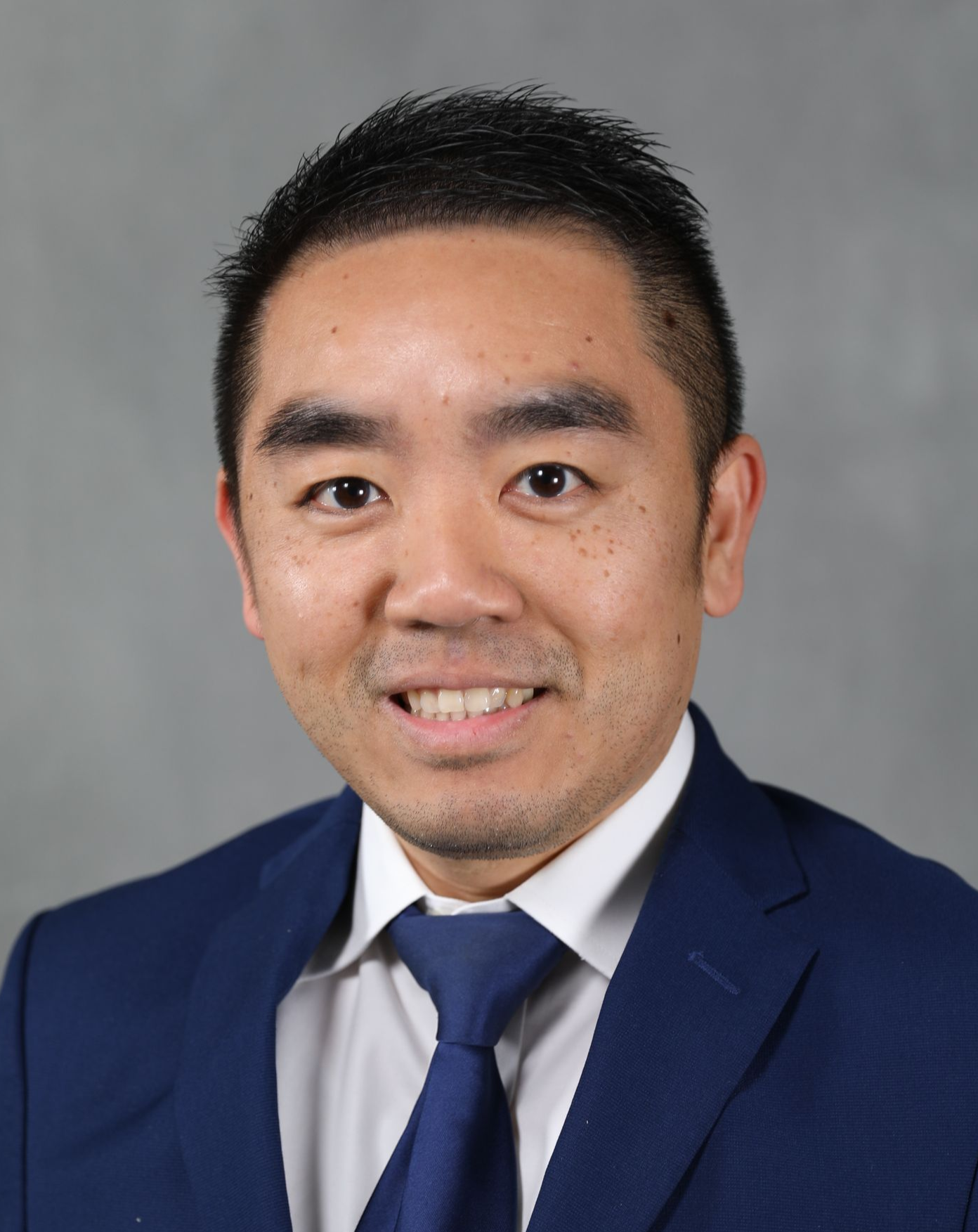 A professional headshot of a person wearing a navy blue suit, white collared shirt, and blue tie against a gray background.