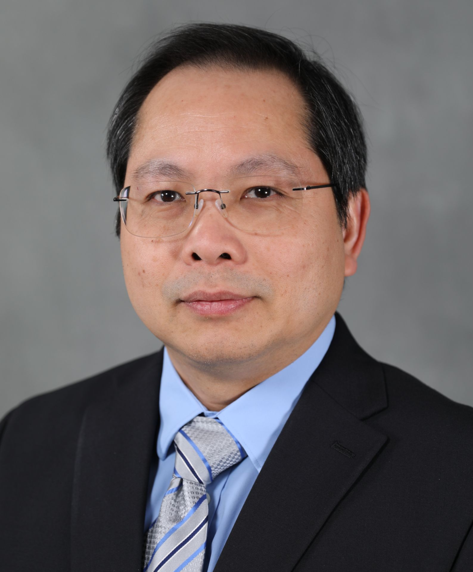 Professional headshot of a person wearing a dark suit, light blue dress shirt, and patterned tie against a gray background.