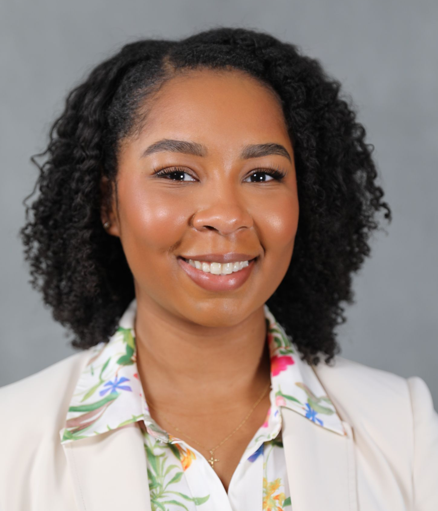A smiling professional with shoulder-length curly hair wearing a white blazer over a floral collared shirt.
