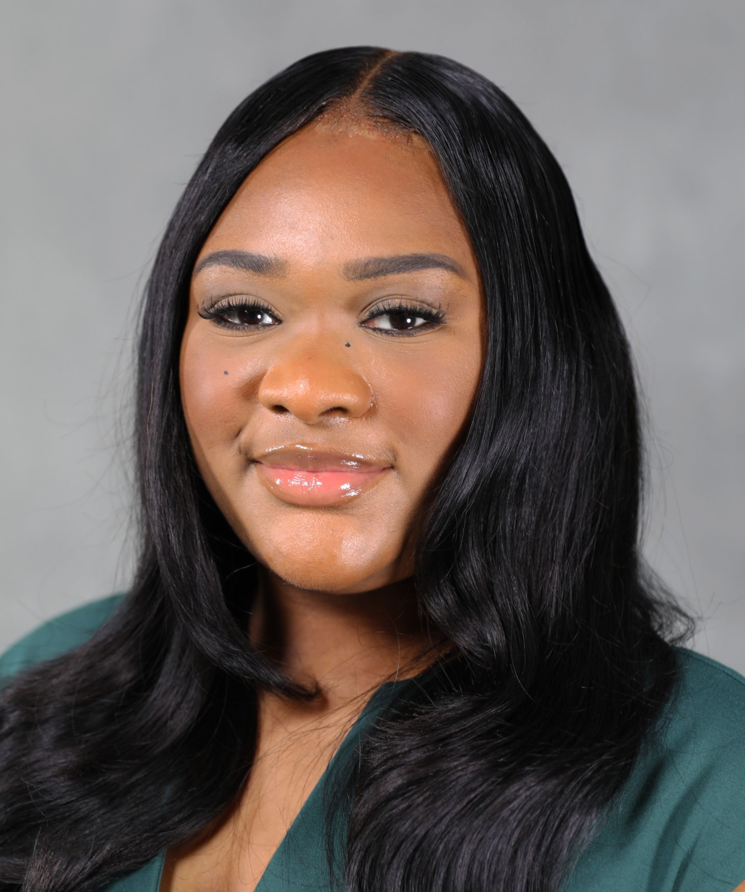 A headshot of a person with long, dark, wavy hair, wearing a dark green top against a neutral gray background.