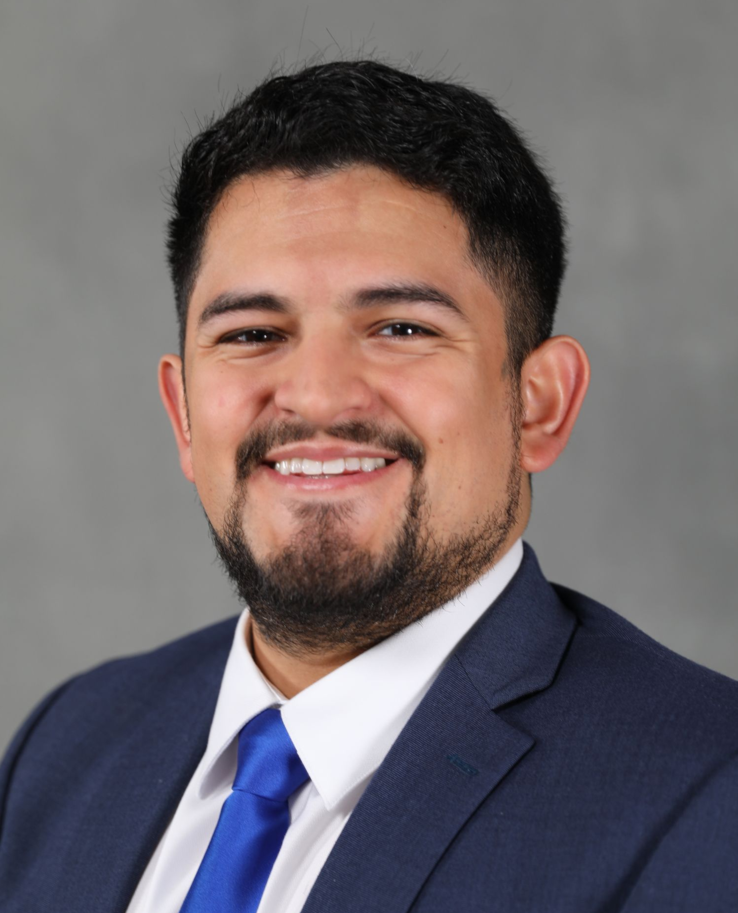 A smiling man with a goatee wearing a blue suit, white shirt, and blue tie against a plain gray background.