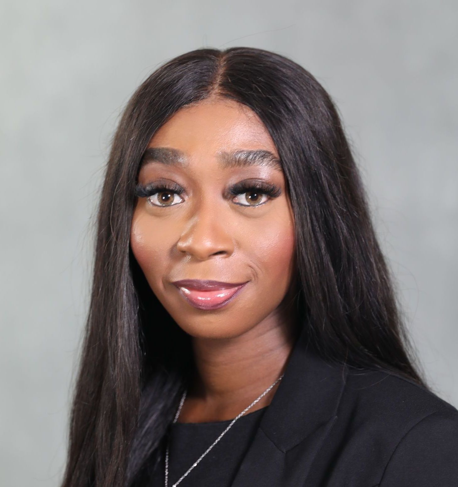 A professional headshot of a person with long, dark hair wearing a black blazer against a neutral grey background.